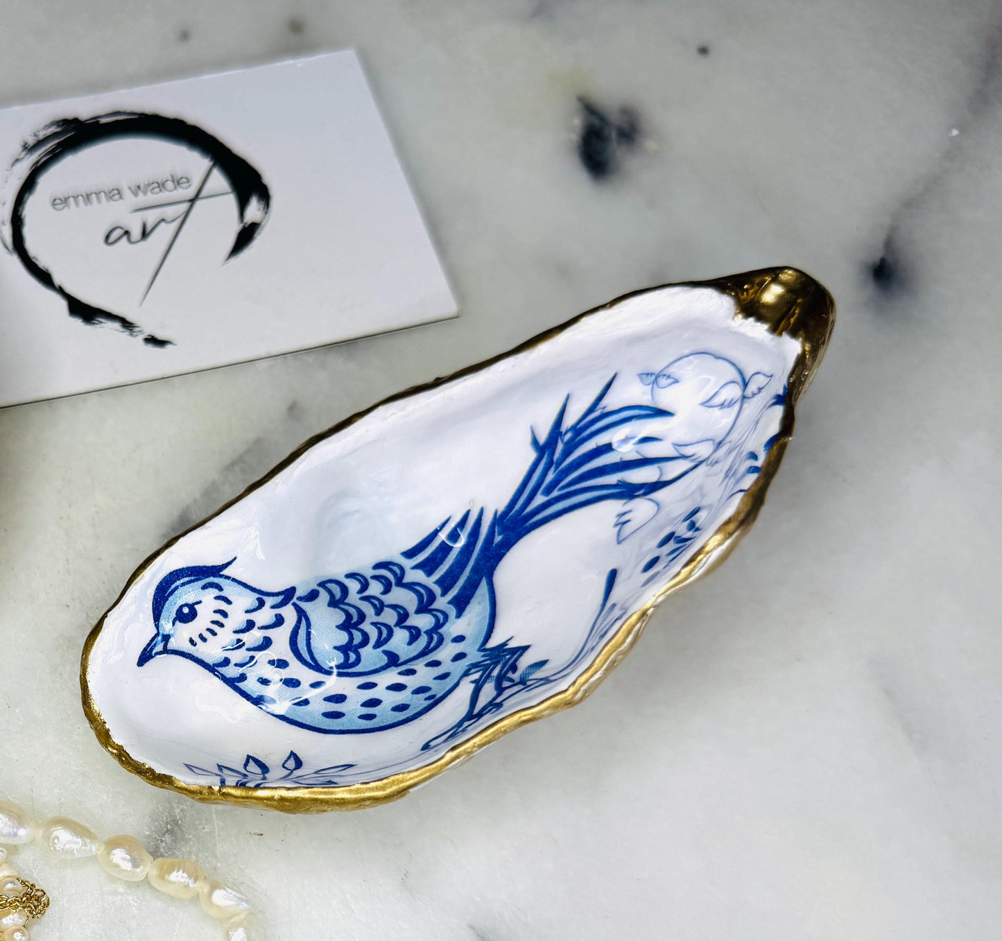 Handmade Dorset oyster shell trinket dish with blue porcelain-style bird design, gold leaf gilded edges and glossy resin finish. Coastal jewellery holder and ring dish photographed on a light surface, showing the natural shell shape and pearly interior. Unique sustainably sourced shell, sold individually as decorative coastal home décor or a thoughtful gift.