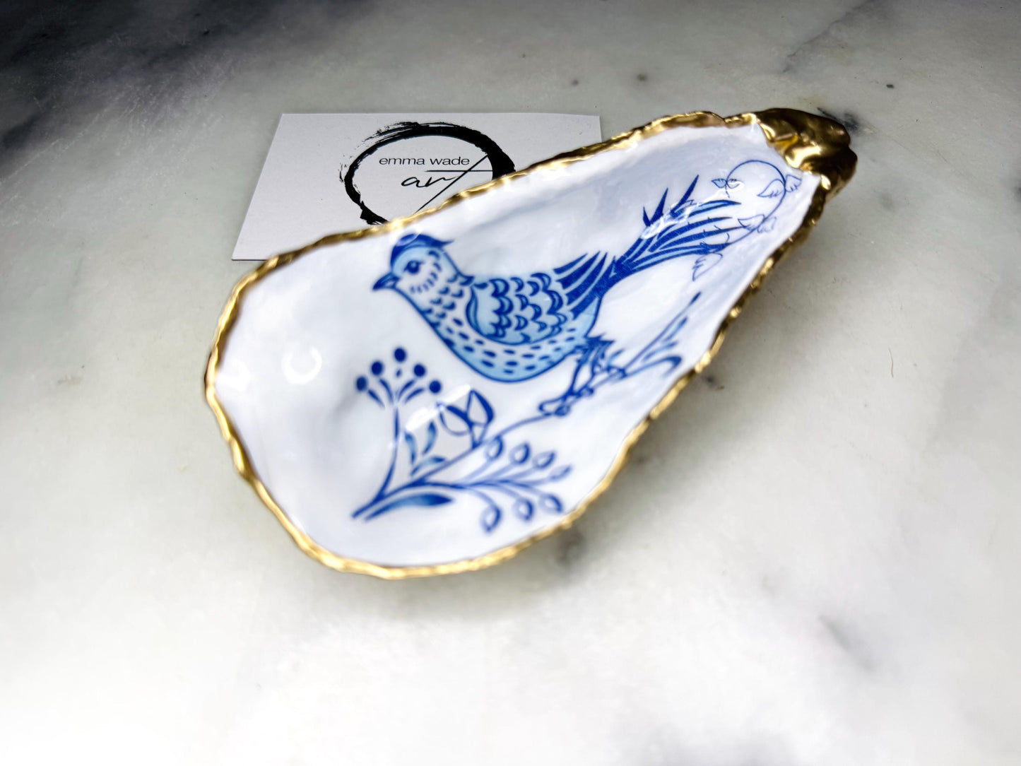 Handmade Dorset oyster shell trinket dish with blue porcelain-style bird design, gold leaf gilded edges and glossy resin finish. Coastal jewellery holder and ring dish photographed on a light surface, showing the natural shell shape and pearly interior. Unique sustainably sourced shell, sold individually as decorative coastal home décor or a thoughtful gift.