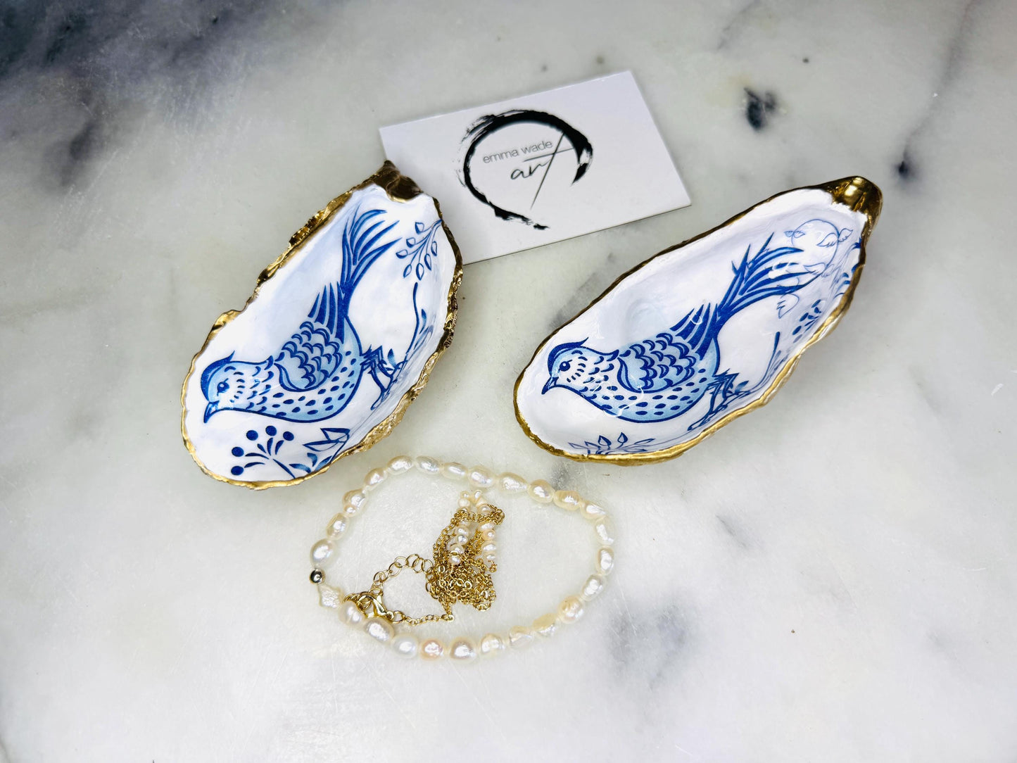 Handmade Dorset oyster shell trinket dish with blue porcelain-style bird design, gold leaf gilded edges and glossy resin finish. Coastal jewellery holder and ring dish photographed on a light surface, showing the natural shell shape and pearly interior. Unique sustainably sourced shell, sold individually as decorative coastal home décor or a thoughtful gift.