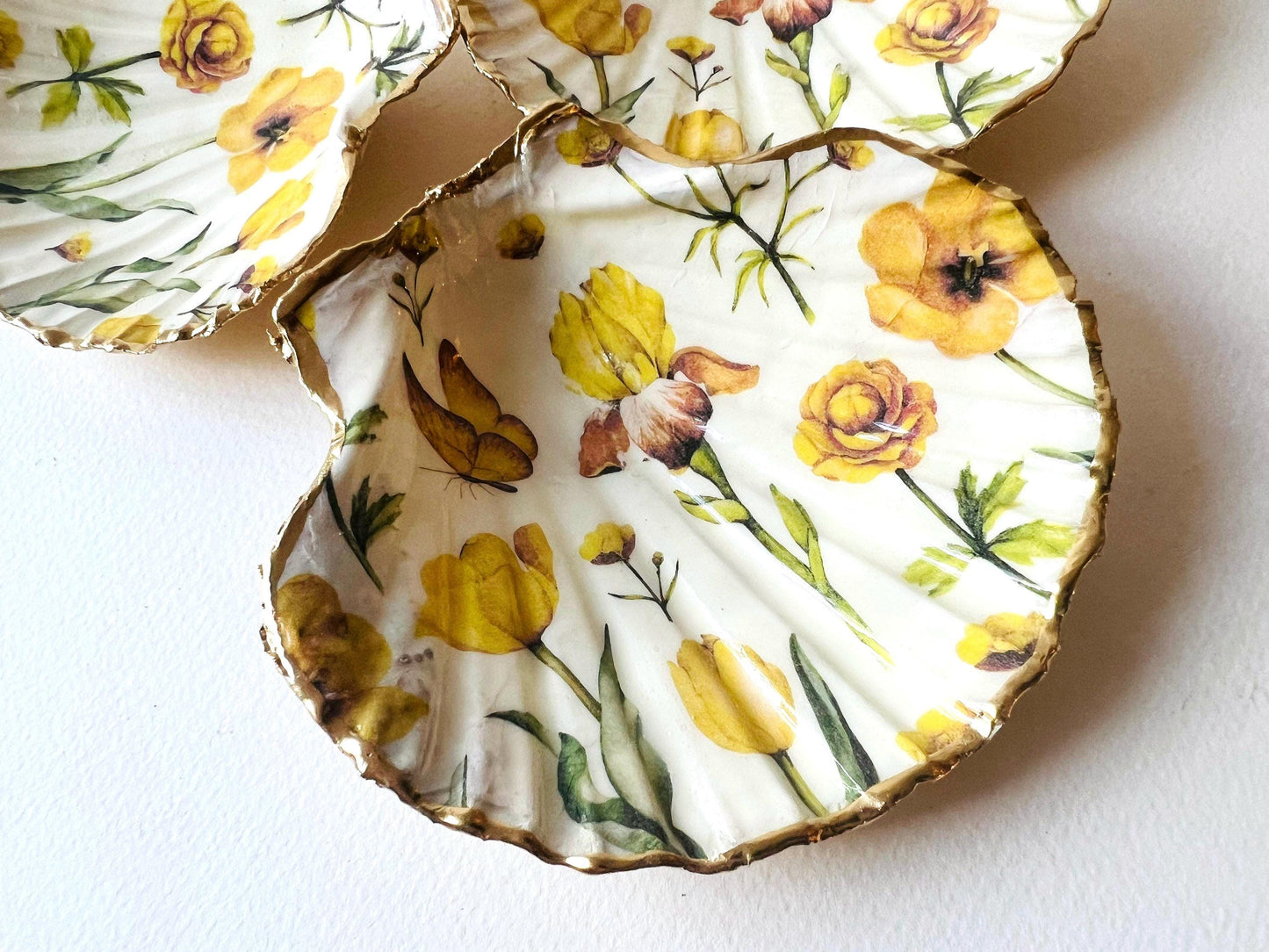 Hand finished scallop shell trinket dish featuring yellow spring flower artwork with a hand gilded gold leaf rim, sealed in glossy clear resin for durability and shine. Coastal inspired shell jewellery dish photographed on a light surface, ideal for rings, earrings and small keepsakes. Decorative floral shell dish by EmmaWadeART inspired by the Dorset coastline, perfect as a gift or elegant home décor accent.