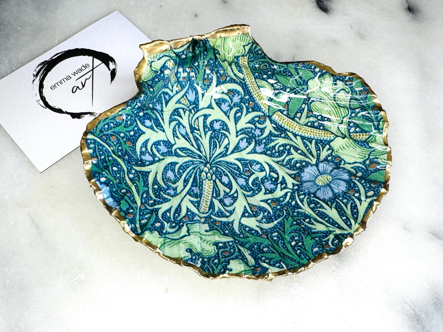 Handmade William Morris inspired scallop shell trinket dish in green and blue tones, featuring a seaweed decoupage design. Finished with gold leaf and sealed with glossy resin. Coastal jewellery dish made from a UK scallop shell, perfect for holding rings, earrings and small treasures. Decorative shell bowl photographed on a light background, showing the natural shell shape and gilded edges.
