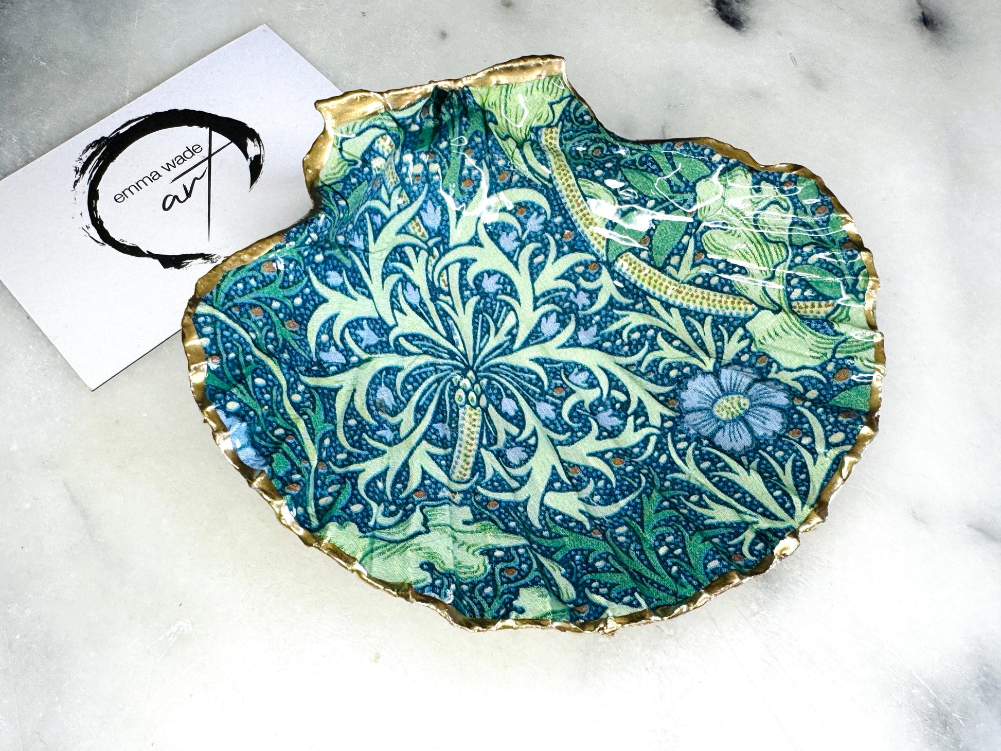 Handmade William Morris inspired scallop shell trinket dish in green and blue tones, featuring a seaweed decoupage design. Finished with gold leaf and sealed with glossy resin. Coastal jewellery dish made from a UK scallop shell, perfect for holding rings, earrings and small treasures. Decorative shell bowl photographed on a light background, showing the natural shell shape and gilded edges.