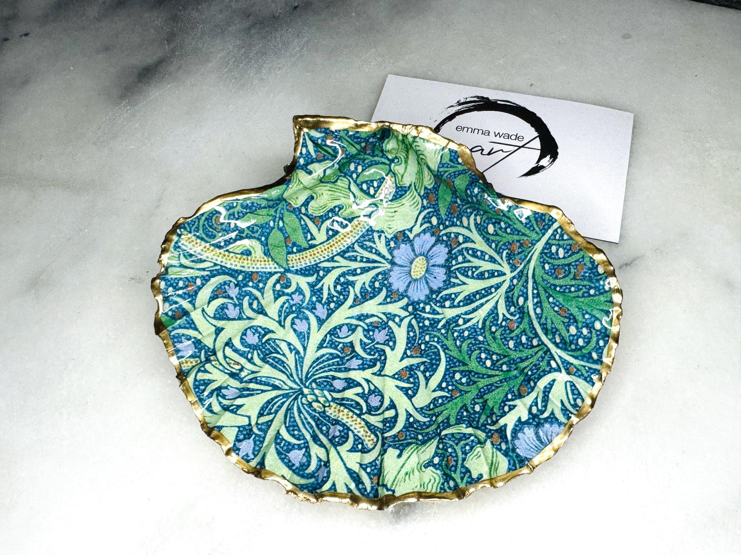 Handmade William Morris inspired scallop shell trinket dish in green and blue tones, featuring a seaweed decoupage design. Finished with gold leaf and sealed with glossy resin. Coastal jewellery dish made from a UK scallop shell, perfect for holding rings, earrings and small treasures. Decorative shell bowl photographed on a light background, showing the natural shell shape and gilded edges.