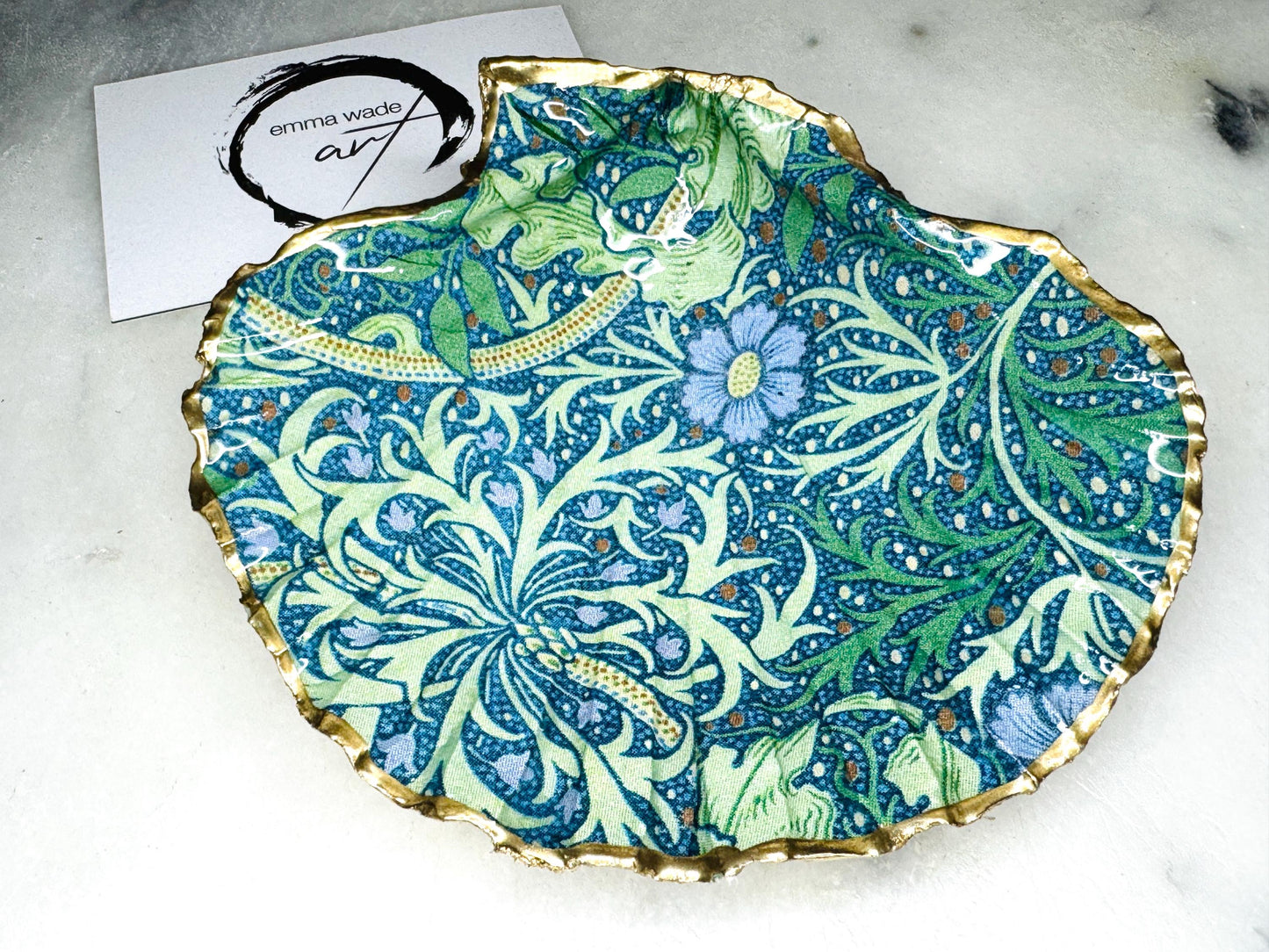 Handmade William Morris inspired scallop shell trinket dish in green and blue tones, featuring a seaweed decoupage design. Finished with gold leaf and sealed with glossy resin. Coastal jewellery dish made from a UK scallop shell, perfect for holding rings, earrings and small treasures. Decorative shell bowl photographed on a light background, showing the natural shell shape and gilded edges.