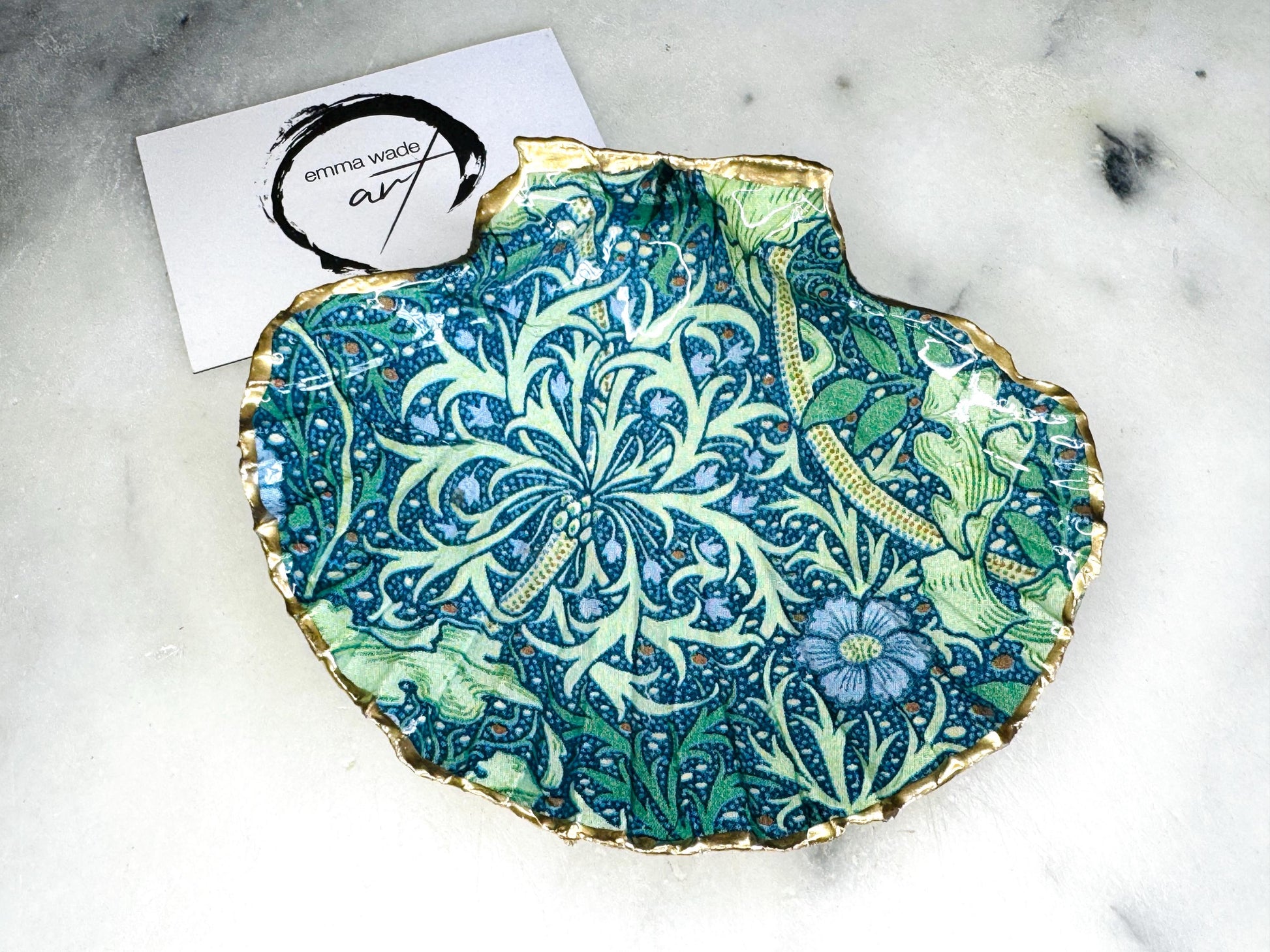 Handmade William Morris inspired scallop shell trinket dish in green and blue tones, featuring a seaweed decoupage design. Finished with gold leaf and sealed with glossy resin. Coastal jewellery dish made from a UK scallop shell, perfect for holding rings, earrings and small treasures. Decorative shell bowl photographed on a light background, showing the natural shell shape and gilded edges.