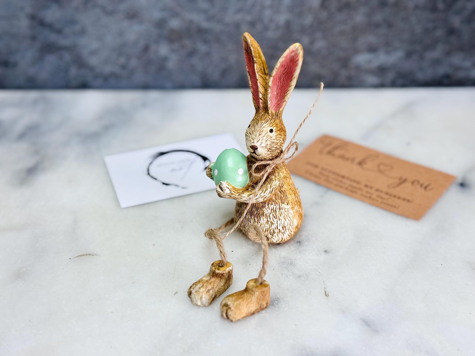 Hand-finished shelf sitting bunny ornament with dangling jute legs and polka dot egg. Available in pink or green. Perfect Easter rabbit shelf decoration, birthday gift, or home décor accessory. Cute animal figurine for rabbit lovers, Easter decorations, new home presents, or seasonal mantel styling.