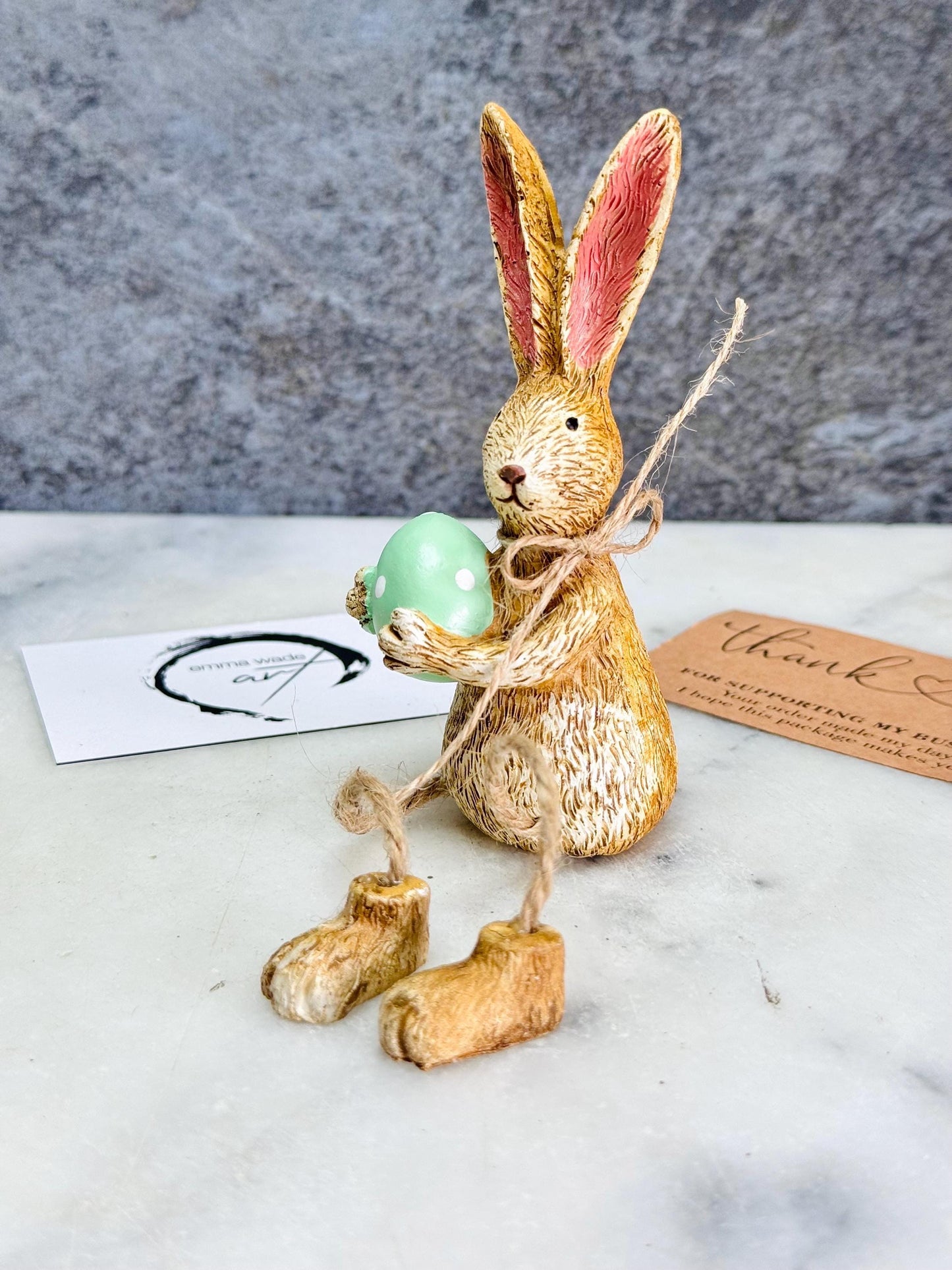 Hand-finished shelf sitting bunny ornament with dangling jute legs and polka dot egg. Available in pink or green. Perfect Easter rabbit shelf decoration, birthday gift, or home décor accessory. Cute animal figurine for rabbit lovers, Easter decorations, new home presents, or seasonal mantel styling.
