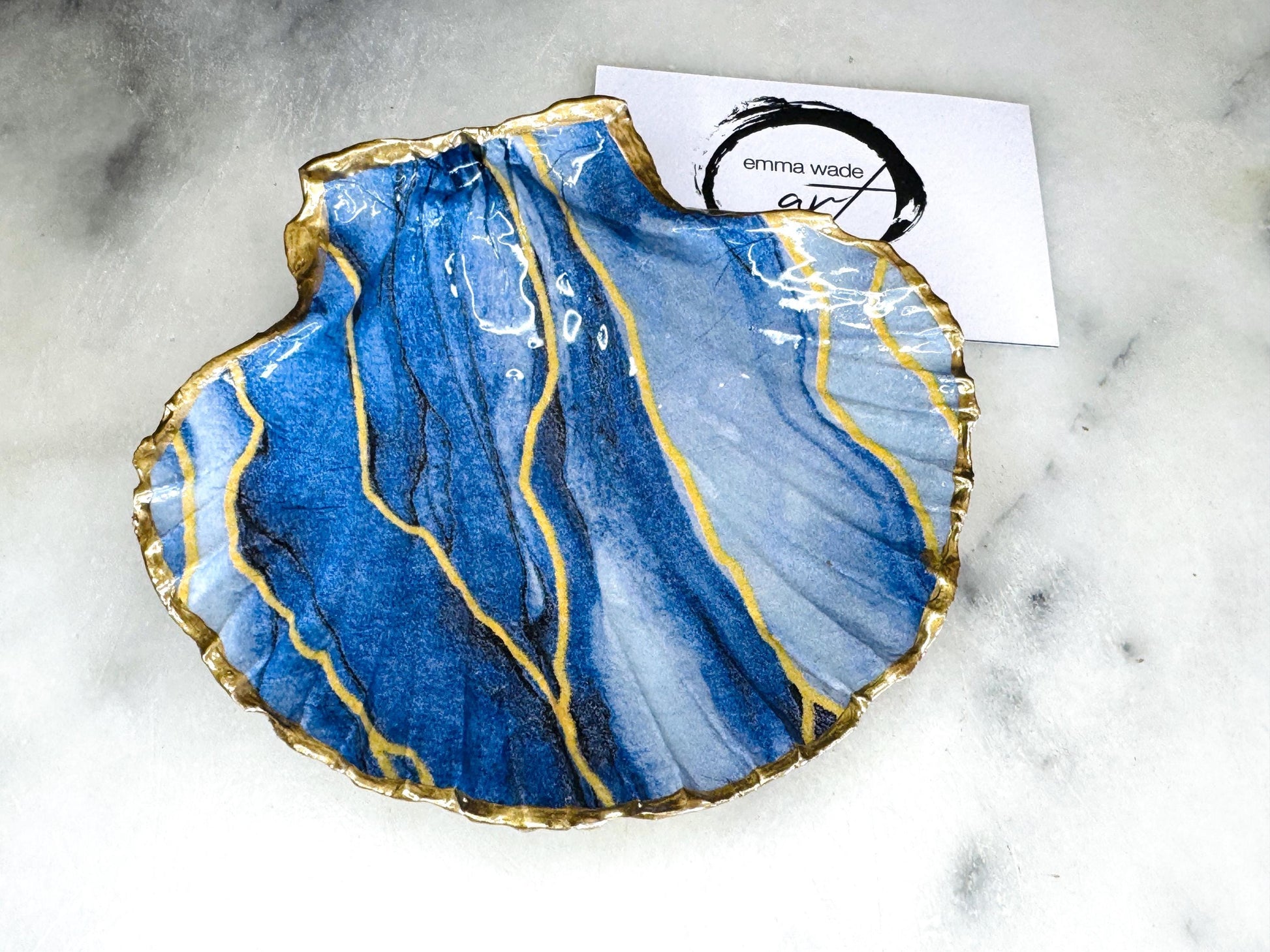 Hand painted blue waves scallop shell trinket dish with gold leaf accents and glossy resin finish, shown with optional gold driftwood display stand. Coastal jewellery and ring dish crafted from a natural Dorset scallop shell, featuring flowing blue wave artwork. Decorative shell bowl photographed on a light background, showing the natural shell shape and gilded edges, ideal for holding rings, earrings, trinkets or small treasures.