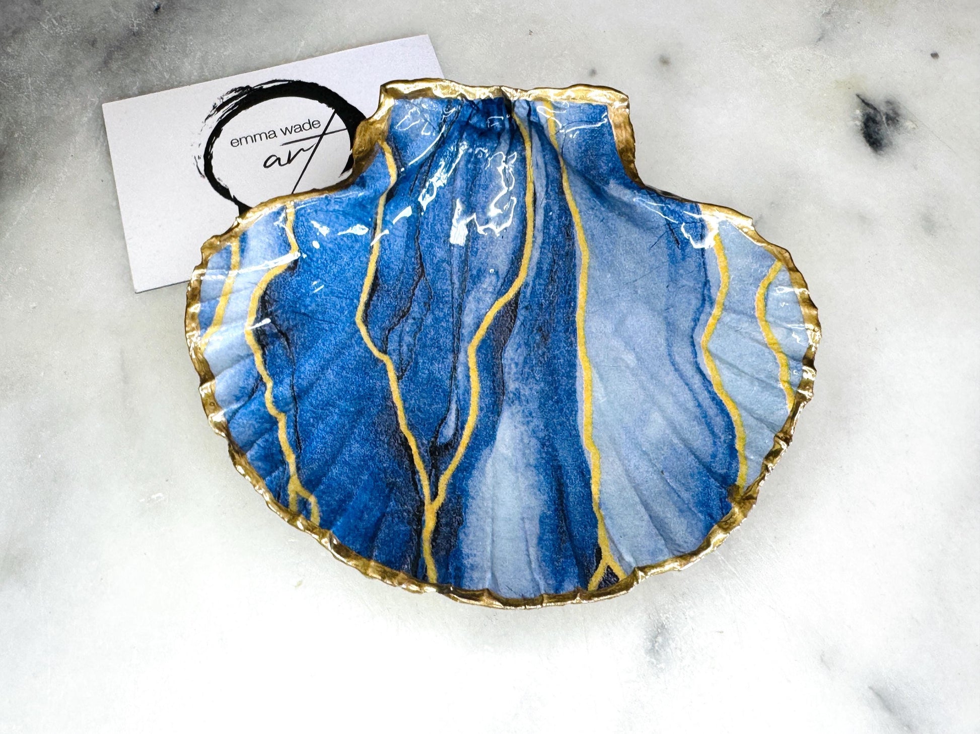 Hand painted blue waves scallop shell trinket dish with gold leaf accents and glossy resin finish, shown with optional gold driftwood display stand. Coastal jewellery and ring dish crafted from a natural Dorset scallop shell, featuring flowing blue wave artwork. Decorative shell bowl photographed on a light background, showing the natural shell shape and gilded edges, ideal for holding rings, earrings, trinkets or small treasures.