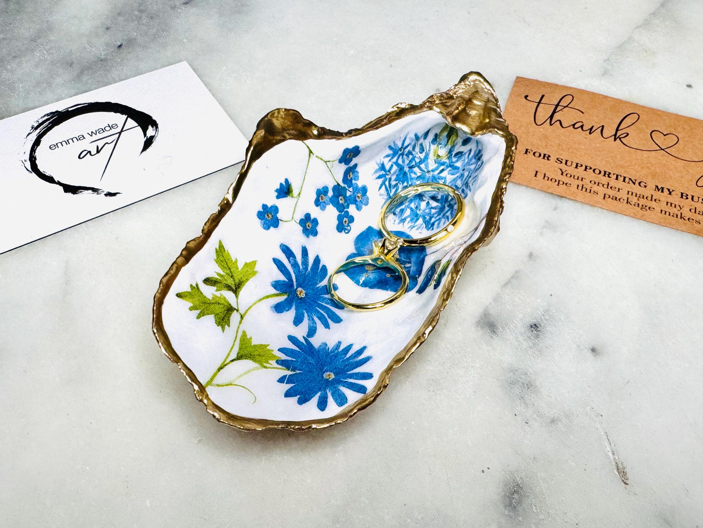 Handmade Dorset oyster shell trinket dish featuring delicate blue floral artwork including bluebells and forget-me-nots, finished with hand-applied gold leaf edging and glossy resin. Coastal-inspired jewellery dish and ring holder available in multiple sizes including XL oyster shells. Elegant handmade home décor gift ideal for birthdays, bridesmaids, Mother’s Day, weddings, and coastal interiors.