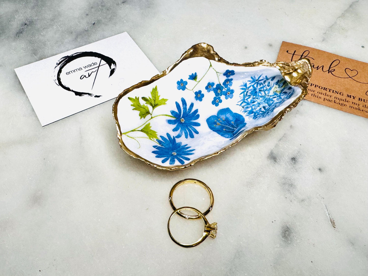 Handmade Dorset oyster shell trinket dish featuring delicate blue floral artwork including bluebells and forget-me-nots, finished with hand-applied gold leaf edging and glossy resin. Coastal-inspired jewellery dish and ring holder available in multiple sizes including XL oyster shells. Elegant handmade home décor gift ideal for birthdays, bridesmaids, Mother’s Day, weddings, and coastal interiors.