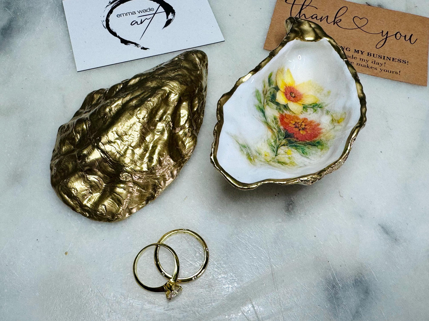 Handmade Dorset oyster shell trinket dish with yellow and orange spring flowers, available with green foliage or stylised blue-toned foliage, finished with a gold leaf rim and glossy resin. Features an original Emma Wade Art design created exclusively for this collection. Shown holding rings on a light background, highlighting the natural shell shape and handcrafted coastal style, ideal as a jewellery dish or keepsake gift.