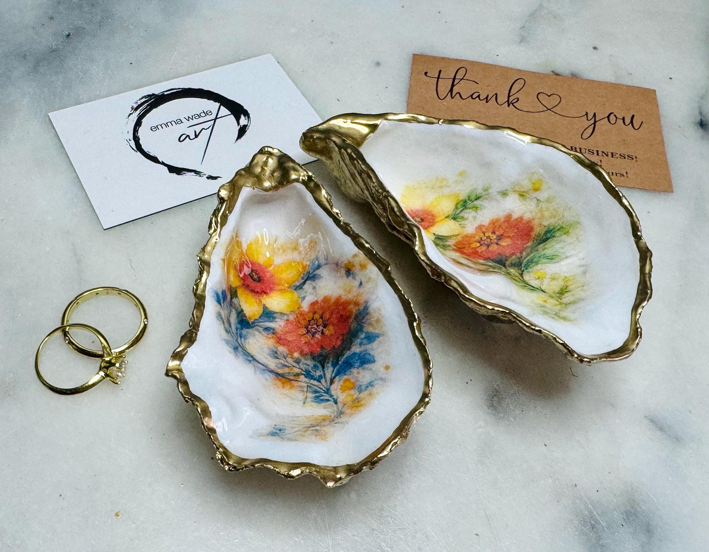 Handmade Dorset oyster shell trinket dish with yellow and orange spring flowers, available with green foliage or stylised blue-toned foliage, finished with a gold leaf rim and glossy resin. Features an original Emma Wade Art design created exclusively for this collection. Shown holding rings on a light background, highlighting the natural shell shape and handcrafted coastal style, ideal as a jewellery dish or keepsake gift.