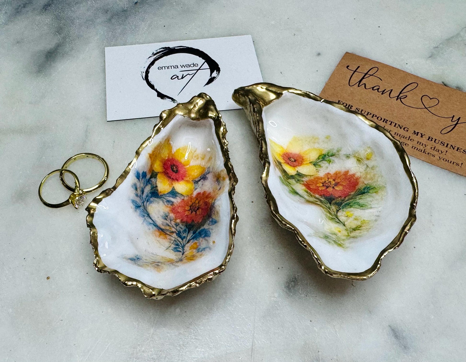Handmade Dorset oyster shell trinket dish with yellow and orange spring flowers, available with green foliage or stylised blue-toned foliage, finished with a gold leaf rim and glossy resin. Features an original Emma Wade Art design created exclusively for this collection. Shown holding rings on a light background, highlighting the natural shell shape and handcrafted coastal style, ideal as a jewellery dish or keepsake gift.