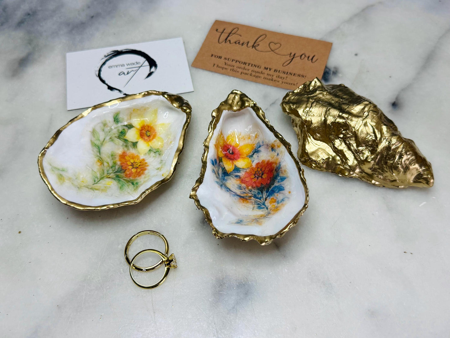 Handmade Dorset oyster shell trinket dish with yellow and orange spring flowers, available with green foliage or stylised blue-toned foliage, finished with a gold leaf rim and glossy resin. Features an original Emma Wade Art design created exclusively for this collection. Shown holding rings on a light background, highlighting the natural shell shape and handcrafted coastal style, ideal as a jewellery dish or keepsake gift.