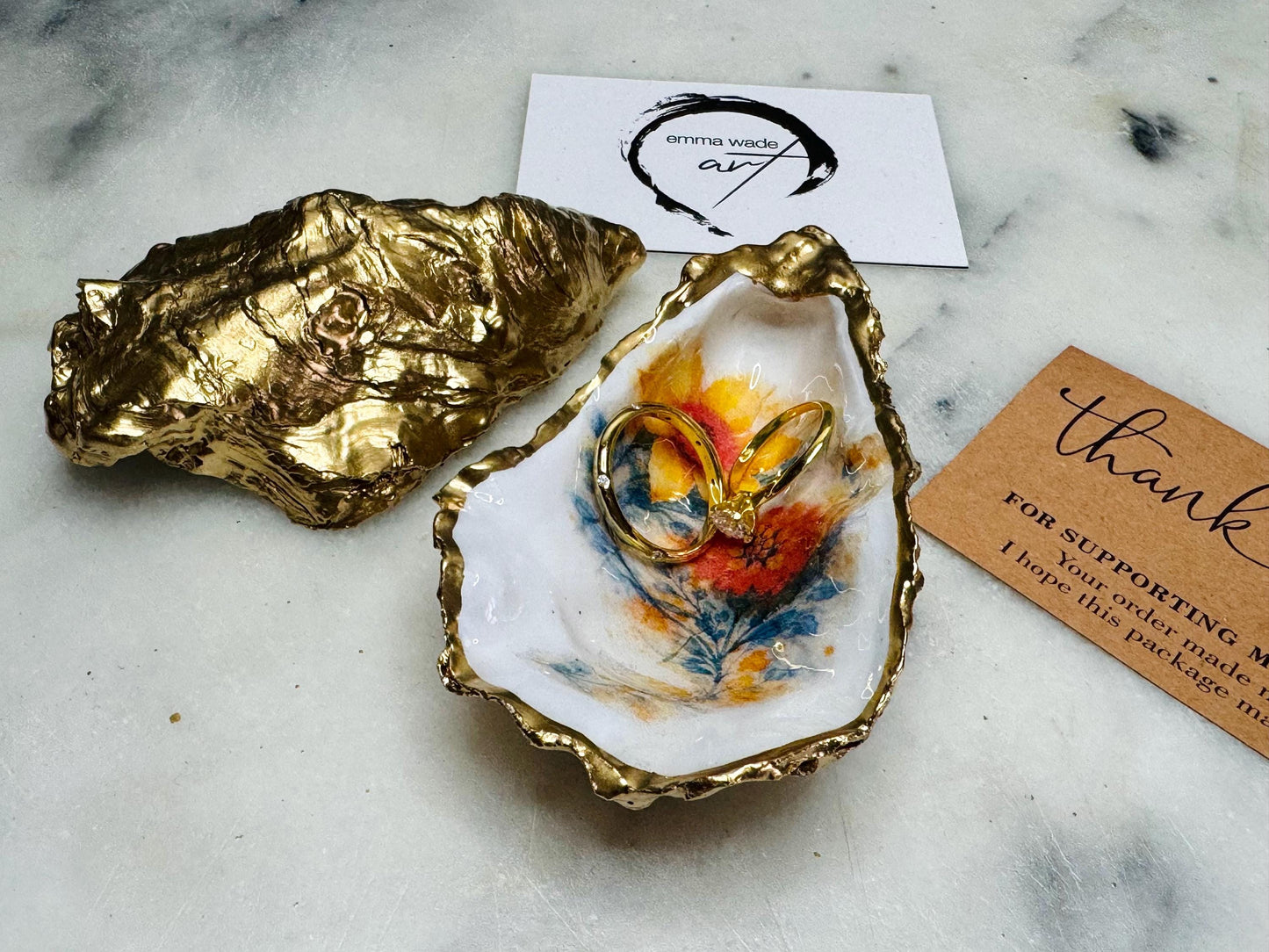 Handmade Dorset oyster shell trinket dish with yellow and orange spring flowers, available with green foliage or stylised blue-toned foliage, finished with a gold leaf rim and glossy resin. Features an original Emma Wade Art design created exclusively for this collection. Shown holding rings on a light background, highlighting the natural shell shape and handcrafted coastal style, ideal as a jewellery dish or keepsake gift.