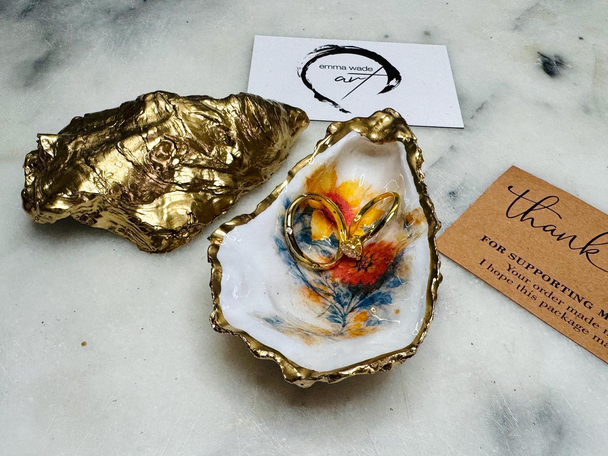 Handmade Dorset oyster shell trinket dish with yellow and orange spring flowers, available with green foliage or stylised blue-toned foliage, finished with a gold leaf rim and glossy resin. Features an original Emma Wade Art design created exclusively for this collection. Shown holding rings on a light background, highlighting the natural shell shape and handcrafted coastal style, ideal as a jewellery dish or keepsake gift.