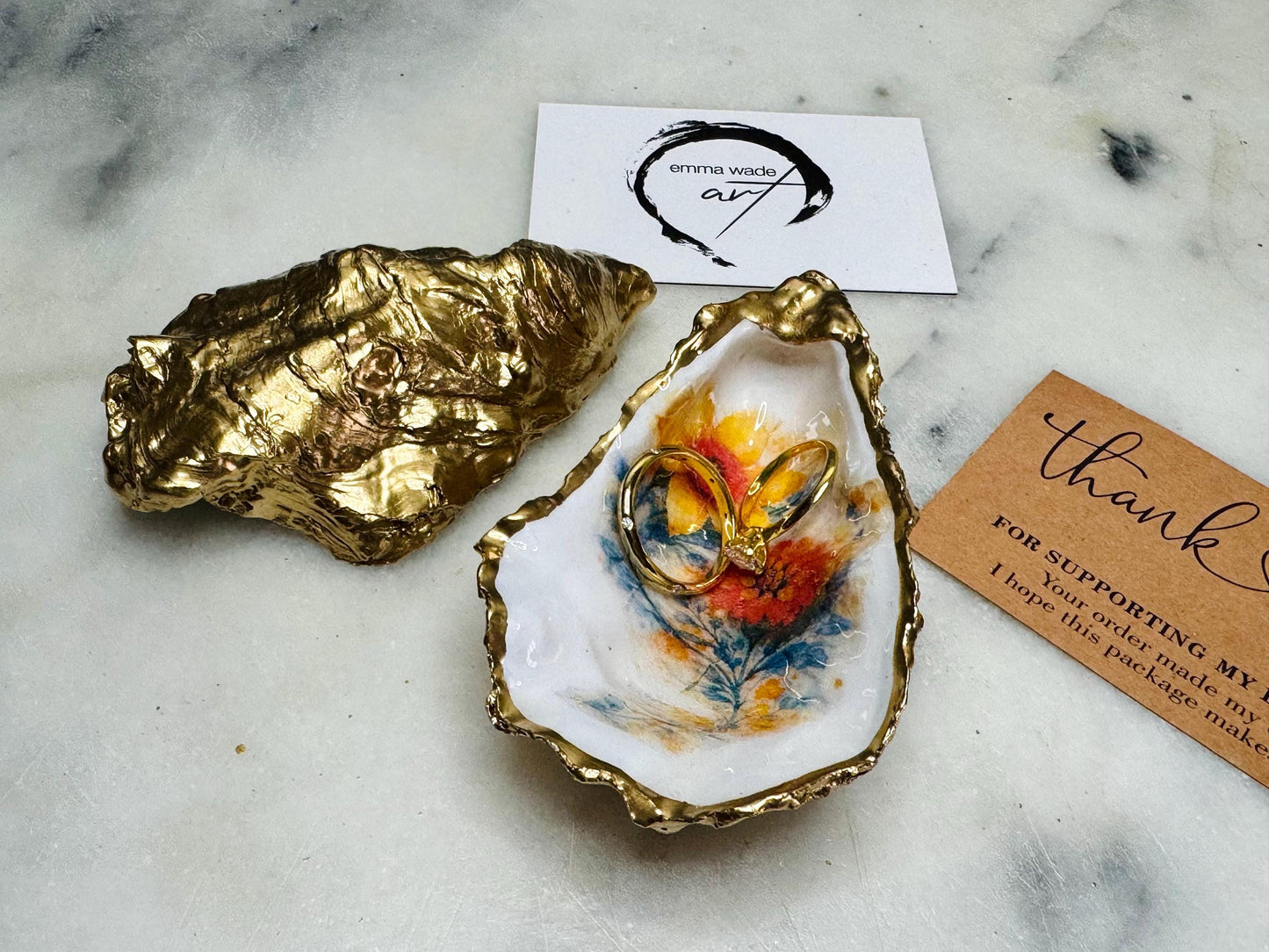 Handmade Dorset oyster shell trinket dish with yellow and orange spring flowers, available with green foliage or stylised blue-toned foliage, finished with a gold leaf rim and glossy resin. Features an original Emma Wade Art design created exclusively for this collection. Shown holding rings on a light background, highlighting the natural shell shape and handcrafted coastal style, ideal as a jewellery dish or keepsake gift.