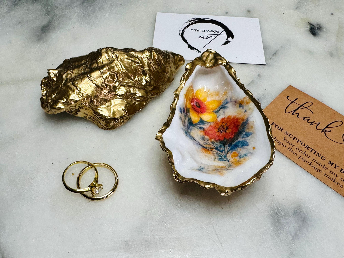 Handmade Dorset oyster shell trinket dish with yellow and orange spring flowers, available with green foliage or stylised blue-toned foliage, finished with a gold leaf rim and glossy resin. Features an original Emma Wade Art design created exclusively for this collection. Shown holding rings on a light background, highlighting the natural shell shape and handcrafted coastal style, ideal as a jewellery dish or keepsake gift.