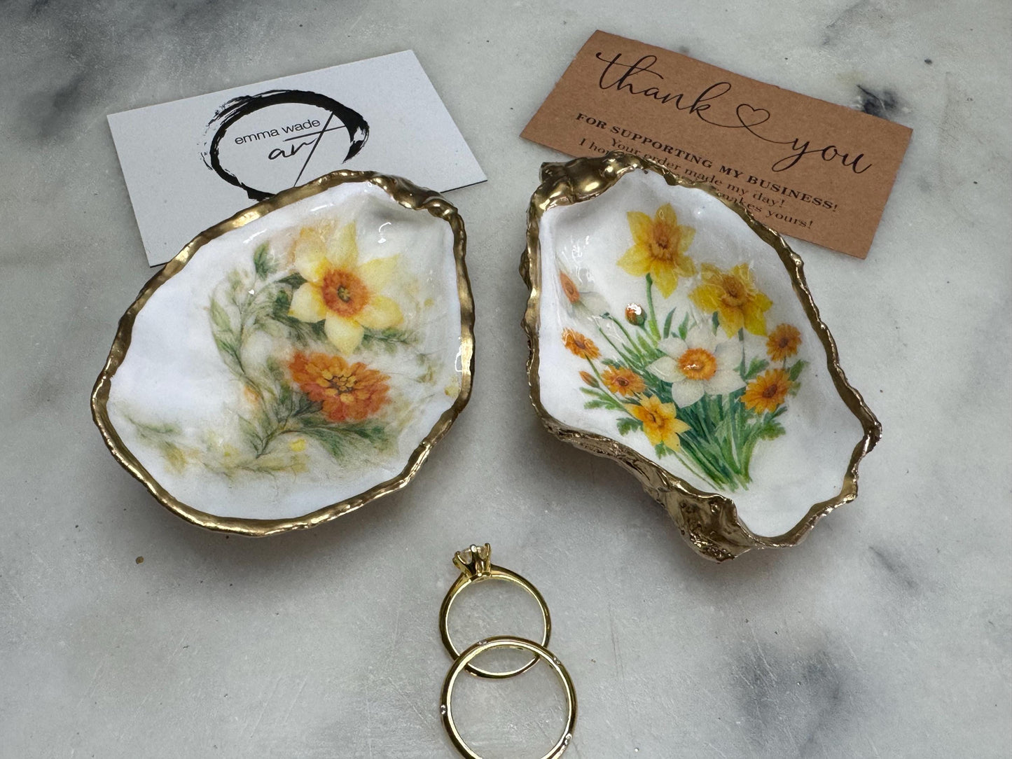 Handmade Dorset oyster shell trinket dish with yellow and orange spring flowers, Daffodils and Marigolds, available in two designs, finished with a gold leaf rim and glossy resin. Features an original Emma Wade Art design created exclusively for this collection. Shown holding rings on a light background, highlighting the natural shell shape and handcrafted coastal style, ideal as a jewellery dish or keepsake gift.