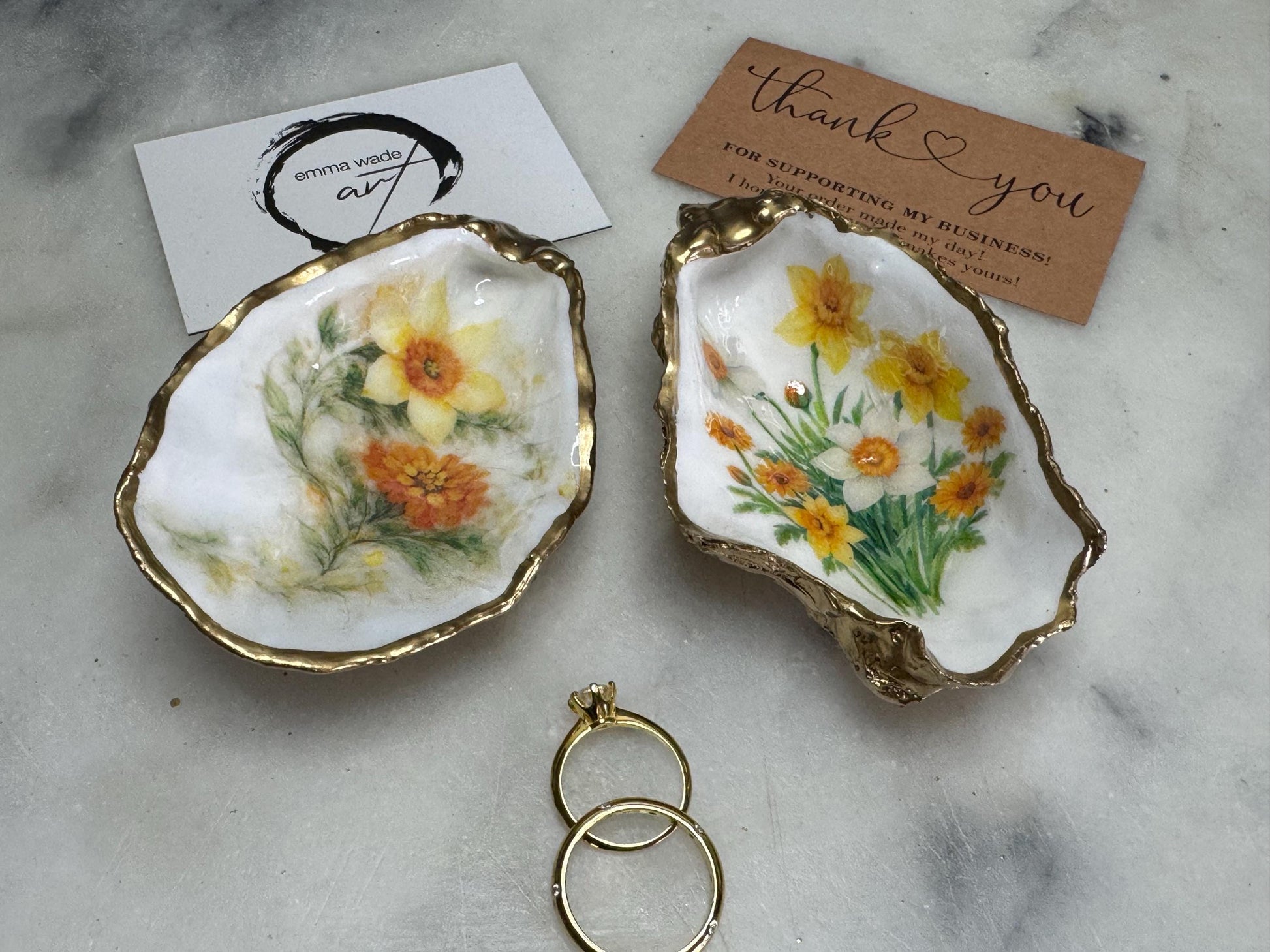 Handmade Dorset oyster shell trinket dish with yellow and orange spring flowers, Daffodils and Marigolds, available in two designs, finished with a gold leaf rim and glossy resin. Features an original Emma Wade Art design created exclusively for this collection. Shown holding rings on a light background, highlighting the natural shell shape and handcrafted coastal style, ideal as a jewellery dish or keepsake gift.