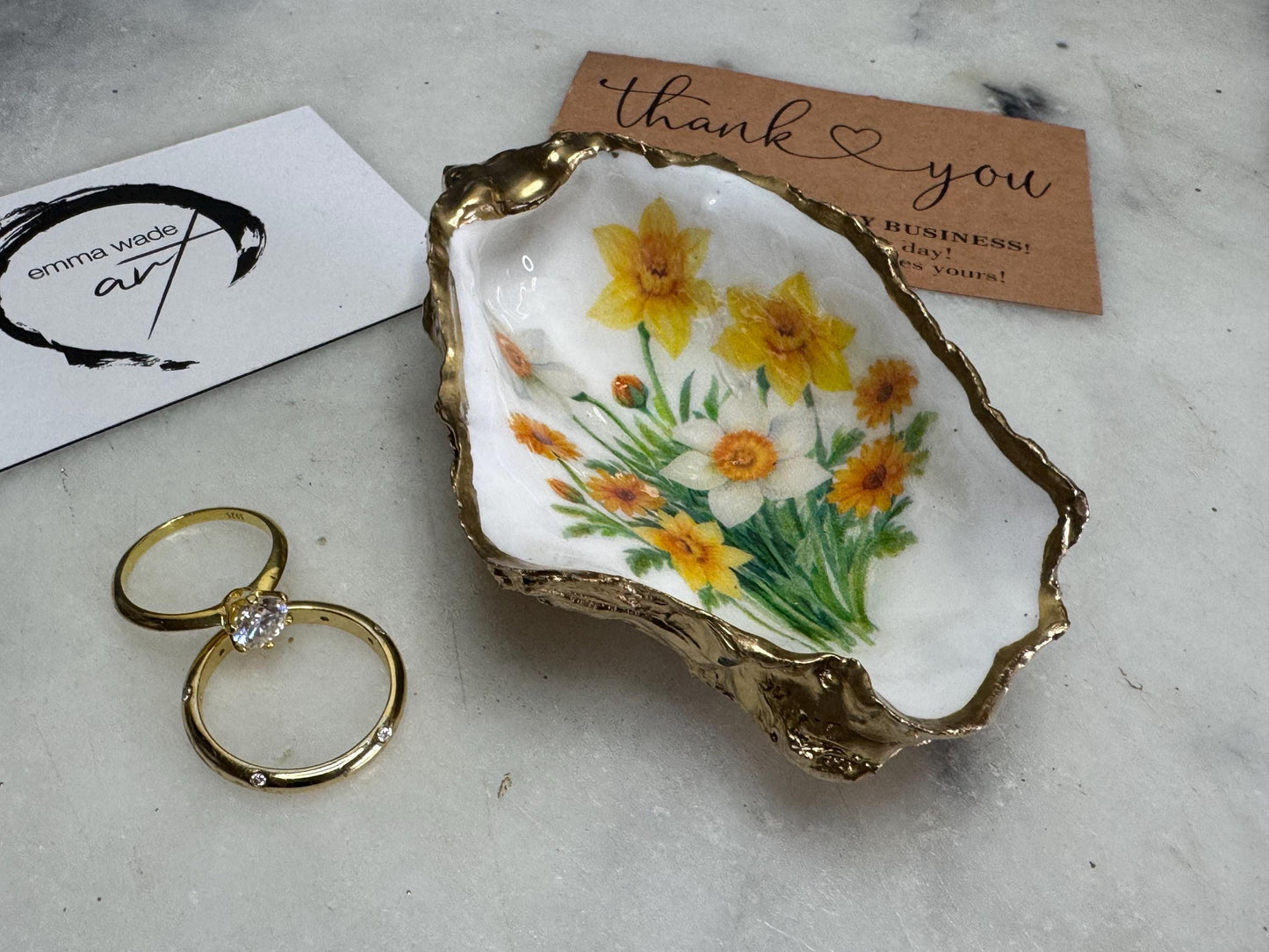 Handmade Dorset oyster shell trinket dish with yellow and orange spring flowers, Daffodils and Marigolds, available in two designs, finished with a gold leaf rim and glossy resin. Features an original Emma Wade Art design created exclusively for this collection. Shown holding rings on a light background, highlighting the natural shell shape and handcrafted coastal style, ideal as a jewellery dish or keepsake gift.
