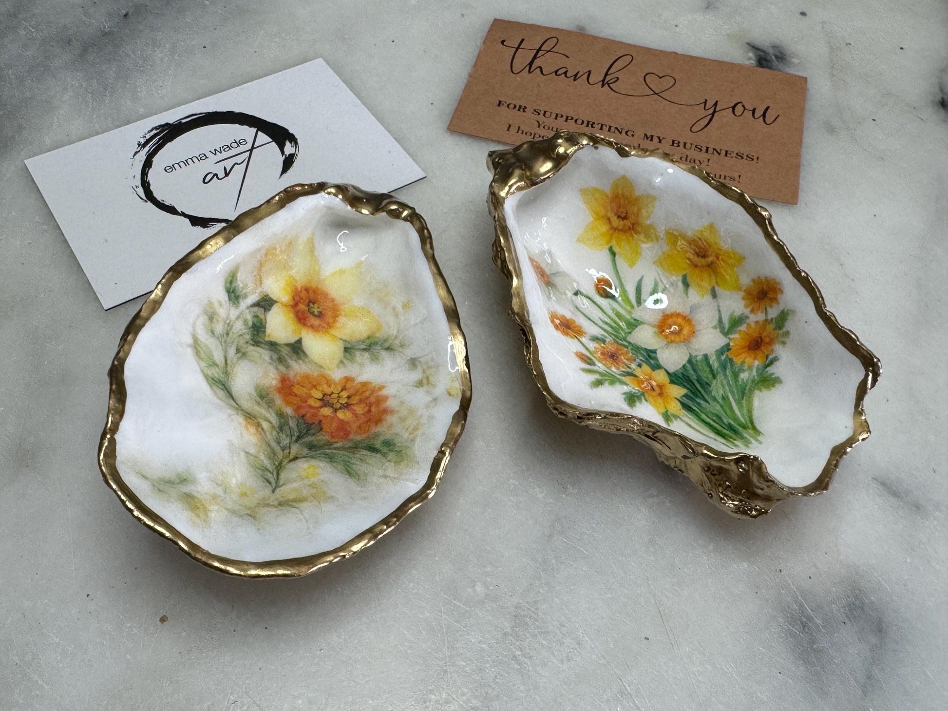 Handmade Dorset oyster shell trinket dish with yellow and orange spring flowers, Daffodils and Marigolds, available in two designs, finished with a gold leaf rim and glossy resin. Features an original Emma Wade Art design created exclusively for this collection. Shown holding rings on a light background, highlighting the natural shell shape and handcrafted coastal style, ideal as a jewellery dish or keepsake gift.