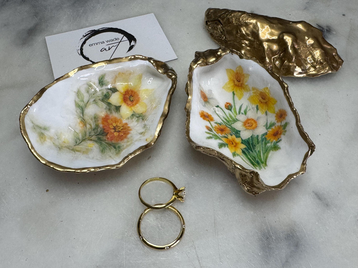 Handmade Dorset oyster shell trinket dish with yellow and orange spring flowers, Daffodils and Marigolds, available in two designs, finished with a gold leaf rim and glossy resin. Features an original Emma Wade Art design created exclusively for this collection. Shown holding rings on a light background, highlighting the natural shell shape and handcrafted coastal style, ideal as a jewellery dish or keepsake gift.