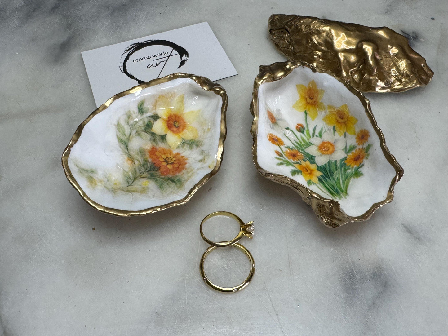 Handmade Dorset oyster shell trinket dish with yellow and orange spring flowers, Daffodils and Marigolds, available in two designs, finished with a gold leaf rim and glossy resin. Features an original Emma Wade Art design created exclusively for this collection. Shown holding rings on a light background, highlighting the natural shell shape and handcrafted coastal style, ideal as a jewellery dish or keepsake gift.