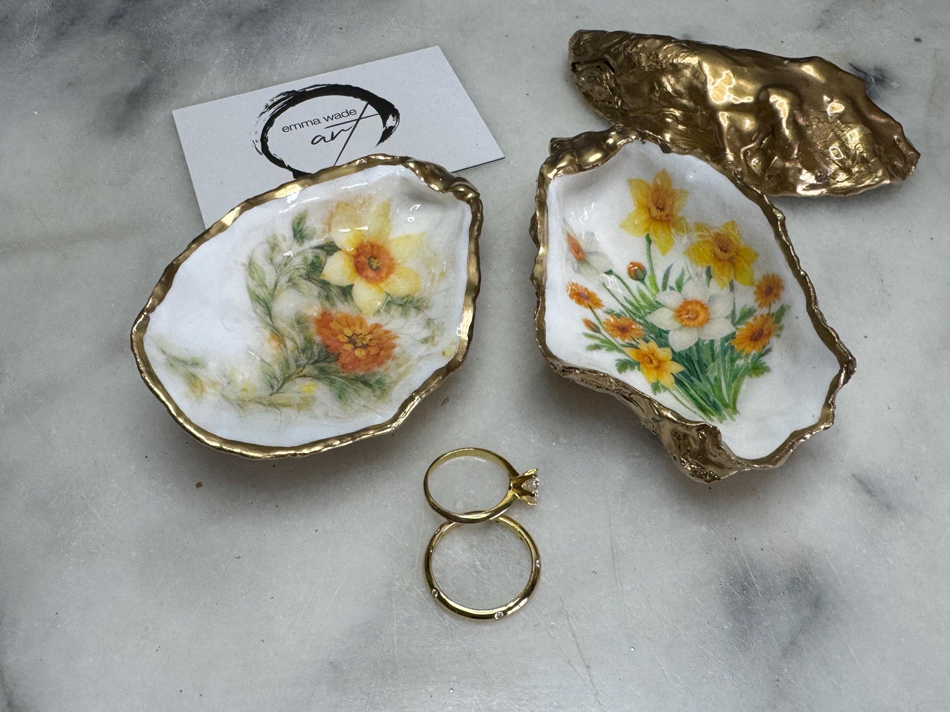 Handmade Dorset oyster shell trinket dish with yellow and orange spring flowers, Daffodils and Marigolds, available in two designs, finished with a gold leaf rim and glossy resin. Features an original Emma Wade Art design created exclusively for this collection. Shown holding rings on a light background, highlighting the natural shell shape and handcrafted coastal style, ideal as a jewellery dish or keepsake gift.