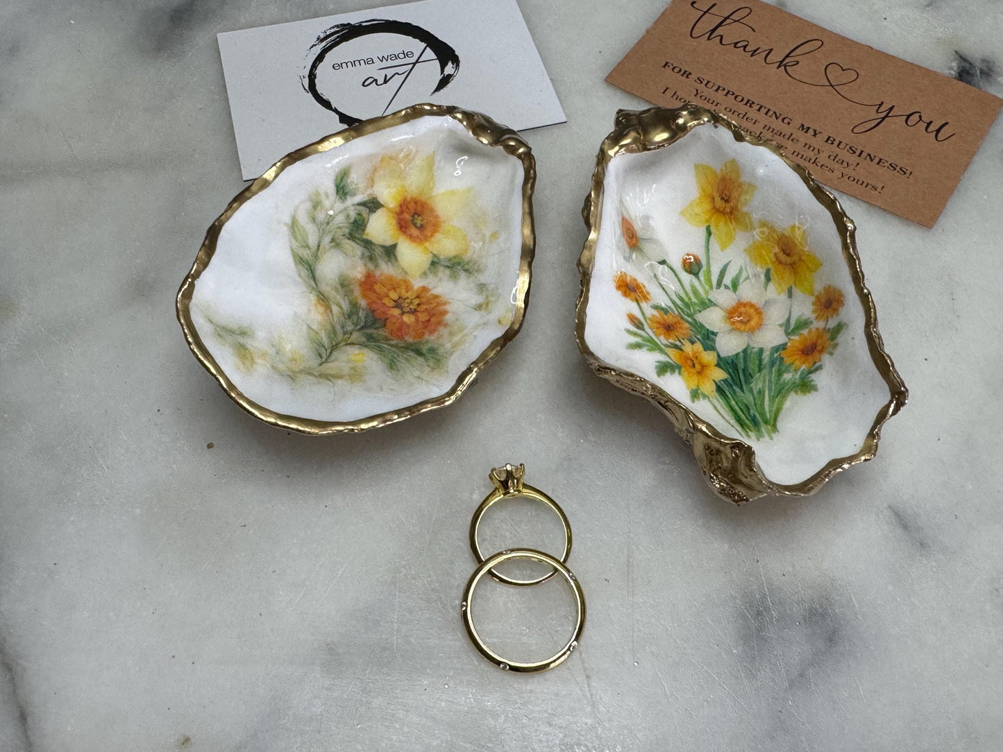 Handmade Dorset oyster shell trinket dish with yellow and orange spring flowers, Daffodils and Marigolds, available in two designs, finished with a gold leaf rim and glossy resin. Features an original Emma Wade Art design created exclusively for this collection. Shown holding rings on a light background, highlighting the natural shell shape and handcrafted coastal style, ideal as a jewellery dish or keepsake gift.