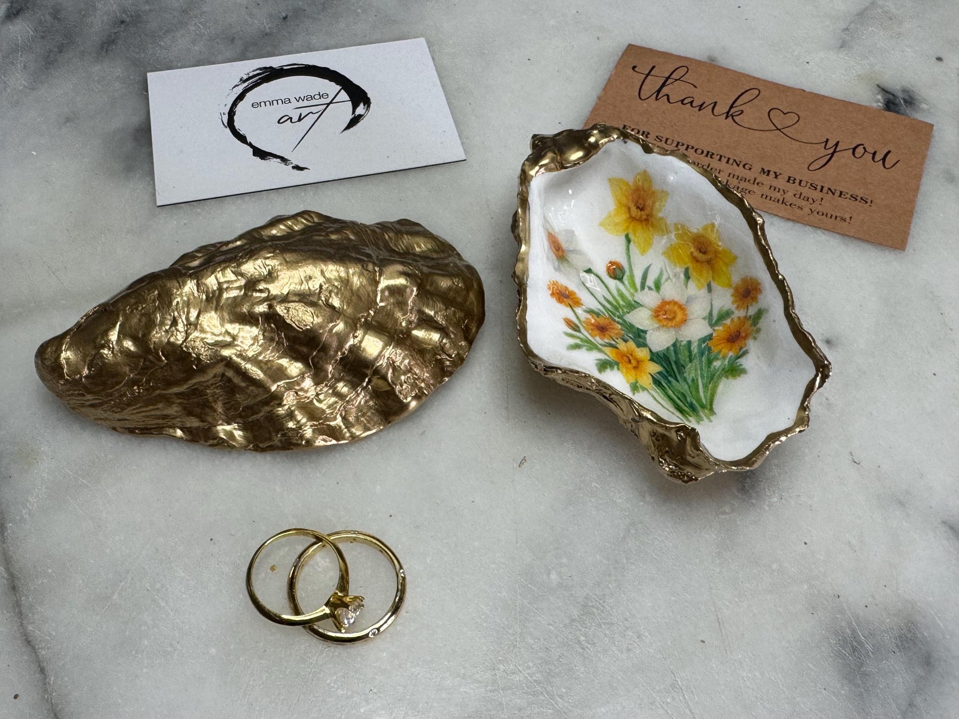 Handmade Dorset oyster shell trinket dish with yellow and orange spring flowers, Daffodils and Marigolds, available in two designs, finished with a gold leaf rim and glossy resin. Features an original Emma Wade Art design created exclusively for this collection. Shown holding rings on a light background, highlighting the natural shell shape and handcrafted coastal style, ideal as a jewellery dish or keepsake gift.