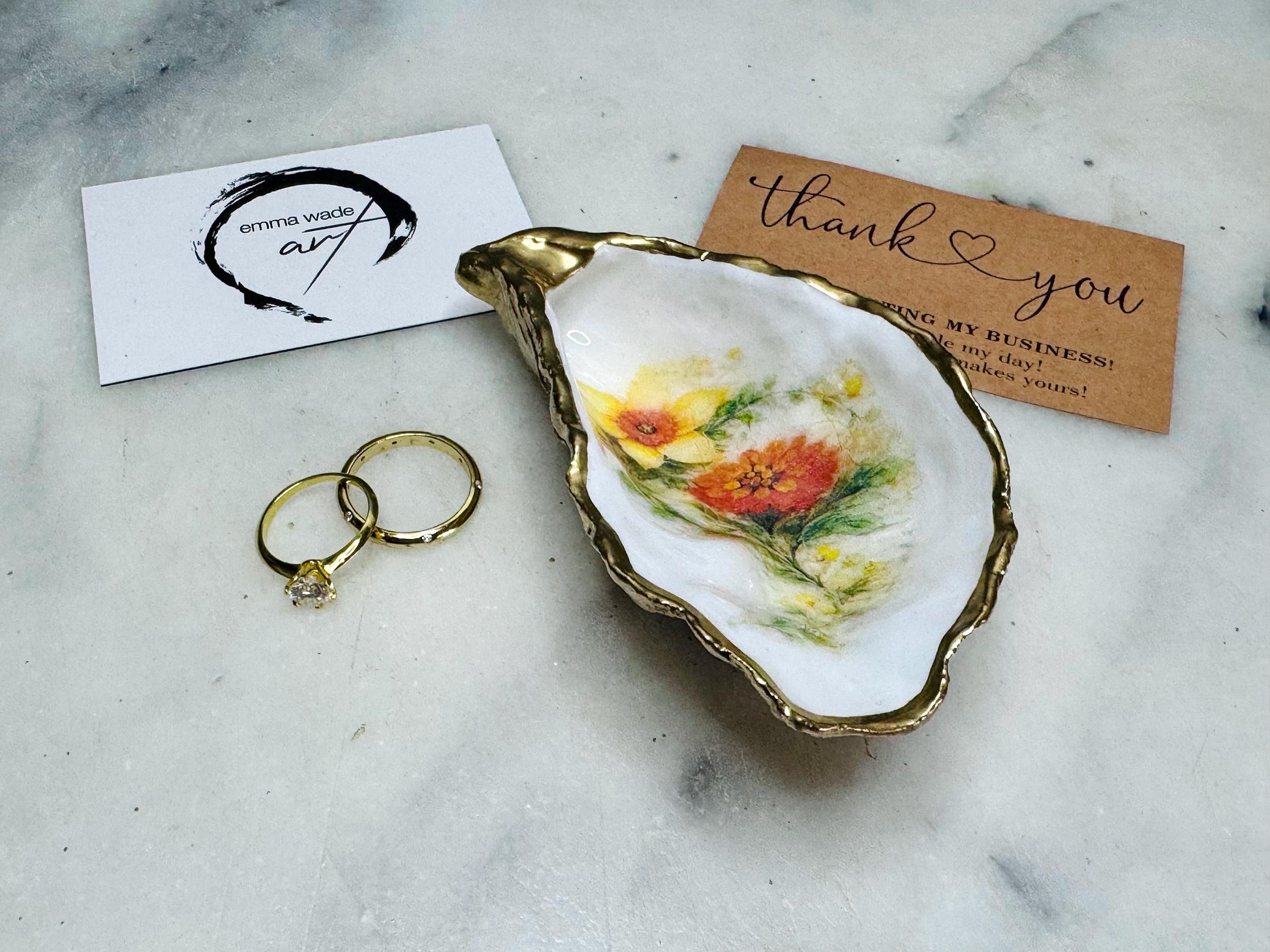 Handmade Dorset oyster shell trinket dish with yellow and orange spring flowers, Daffodils and Marigolds, available in two designs, finished with a gold leaf rim and glossy resin. Features an original Emma Wade Art design created exclusively for this collection. Shown holding rings on a light background, highlighting the natural shell shape and handcrafted coastal style, ideal as a jewellery dish or keepsake gift.