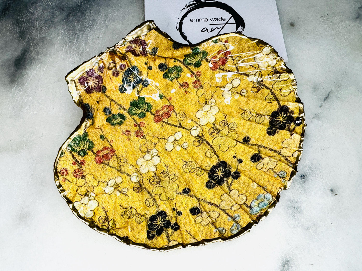 Handcrafted Dorset scallop shell trinket dish featuring a Japanese plum blossom floral design on a rich yellow background, finished with a gold leaf rim and glossy resin. Decorative blossom branches in white, green and black tones, styled on a light marble surface with a gold shell accent. Coastal inspired jewellery tray or ring holder, ideal as a handmade decorative keepsake gift. William Morris Design.
