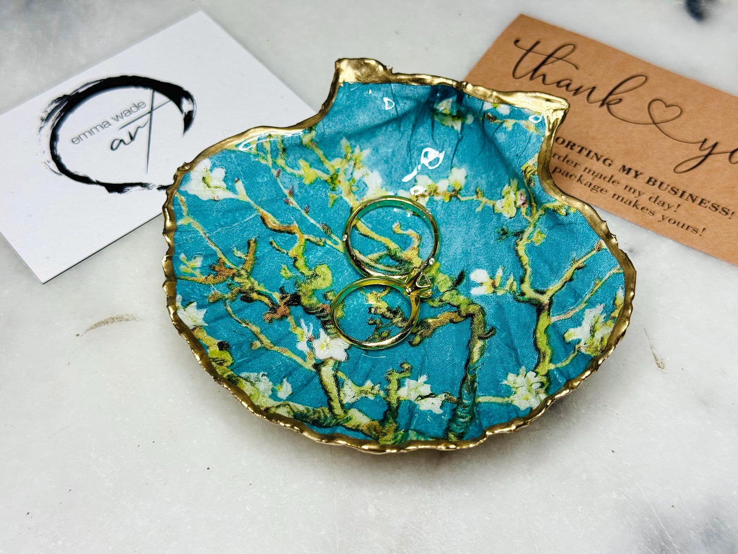 Handcrafted Dorset scallop shell trinket dish inspired by Vincent van Gogh’s Almond Blossom painting. Turquoise blue and green background with branching blossom design and raised gold detailing, finished with liquid gold leaf edging and sealed in high-gloss resin. Decorative shell jewellery holder styled on marble surface, suitable for rings, earrings, trinkets or as a tea light holder. Coastal art-inspired home decor piece.
