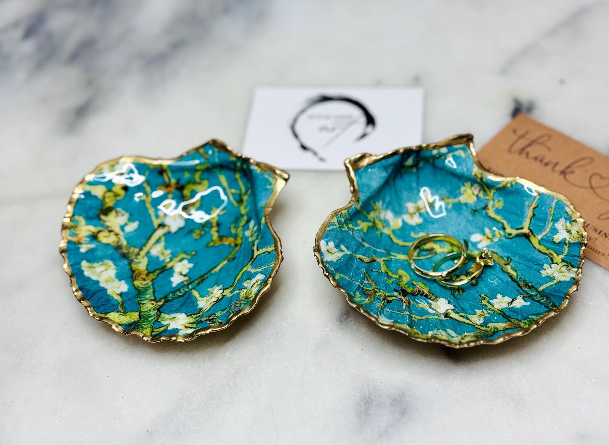 Handcrafted Dorset scallop shell trinket dish inspired by Vincent van Gogh’s Almond Blossom painting. Turquoise blue and green background with branching blossom design and raised gold detailing, finished with liquid gold leaf edging and sealed in high-gloss resin. Decorative shell jewellery holder styled on marble surface, suitable for rings, earrings, trinkets or as a tea light holder. Coastal art-inspired home decor piece.