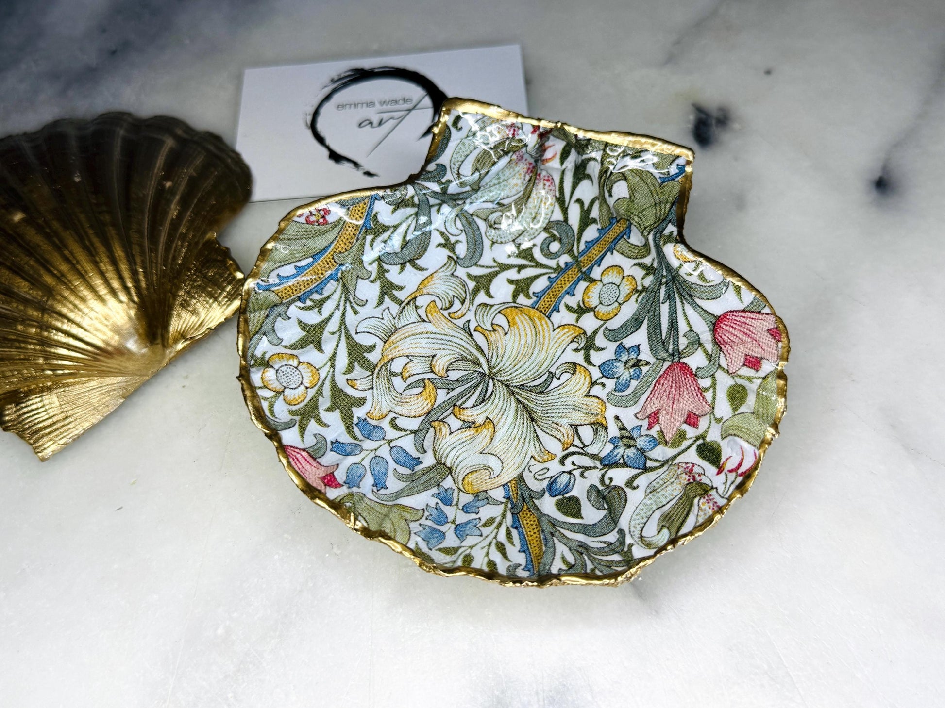 Handcrafted Dorset scallop shell trinket dish featuring William Morris Golden Lily floral design. Natural shell edged with hand applied gold leaf and sealed with glossy resin. Heritage floral jewellery holder displayed with rings on marble surface. Elegant coastal home decor gift.