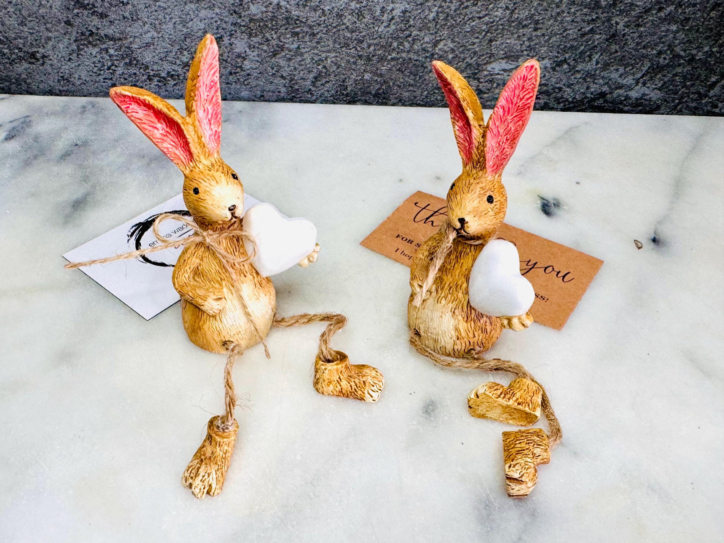 Hand finished rustic bunny shelf sitters holding white wooden hearts, gold and grey rabbit ornaments with pink ears and dangling jute legs, styled on marble surface as charming year-round home decor.