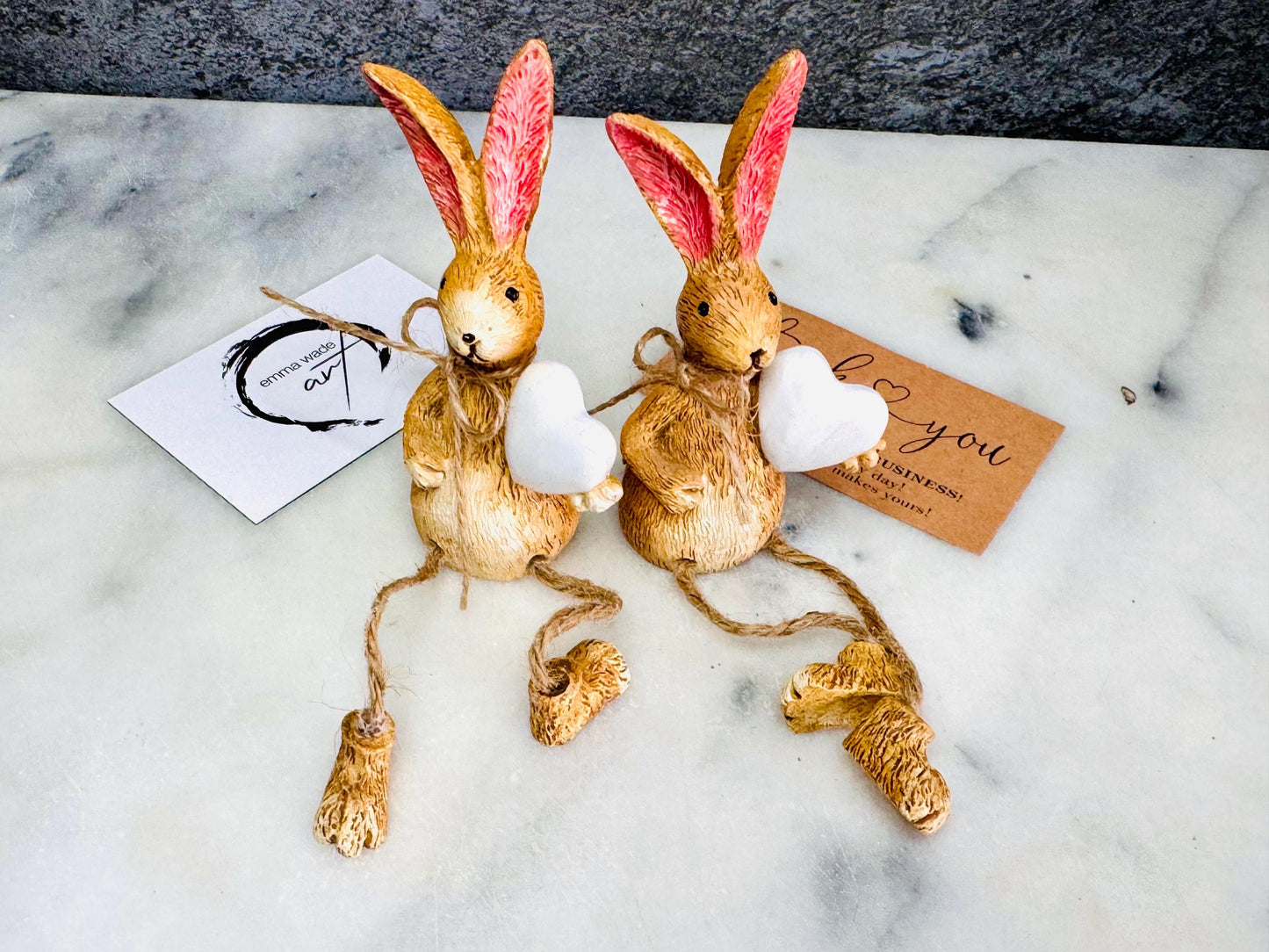 Hand finished rustic bunny shelf sitters holding white wooden hearts, gold and grey rabbit ornaments with pink ears and dangling jute legs, styled on marble surface as charming year-round home decor.