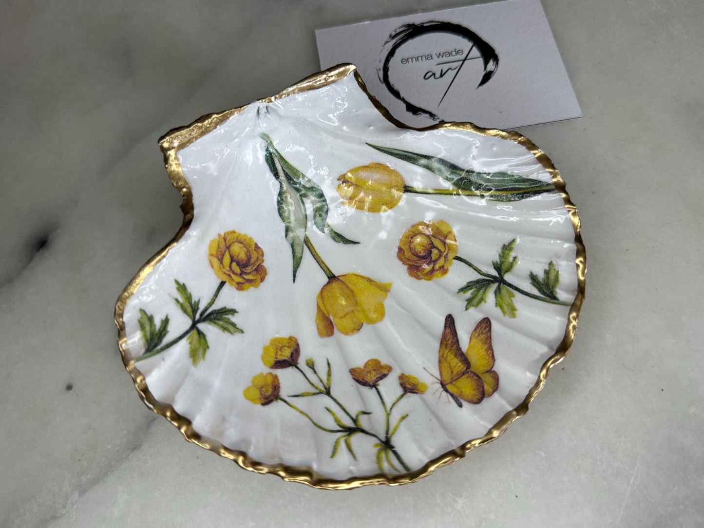 Hand finished scallop shell trinket dish featuring yellow spring flower artwork with a hand gilded gold leaf rim, sealed in glossy clear resin for durability and shine. Coastal inspired shell jewellery dish photographed on a light surface, ideal for rings, earrings and small keepsakes. Decorative floral shell dish by EmmaWadeART inspired by the Dorset coastline, perfect as a gift or elegant home décor accent.