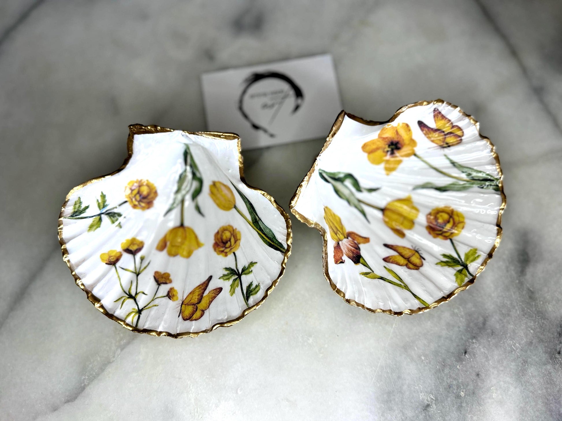 Hand finished scallop shell trinket dish featuring yellow spring flower artwork with a hand gilded gold leaf rim, sealed in glossy clear resin for durability and shine. Coastal inspired shell jewellery dish photographed on a light surface, ideal for rings, earrings and small keepsakes. Decorative floral shell dish by EmmaWadeART inspired by the Dorset coastline, perfect as a gift or elegant home décor accent.