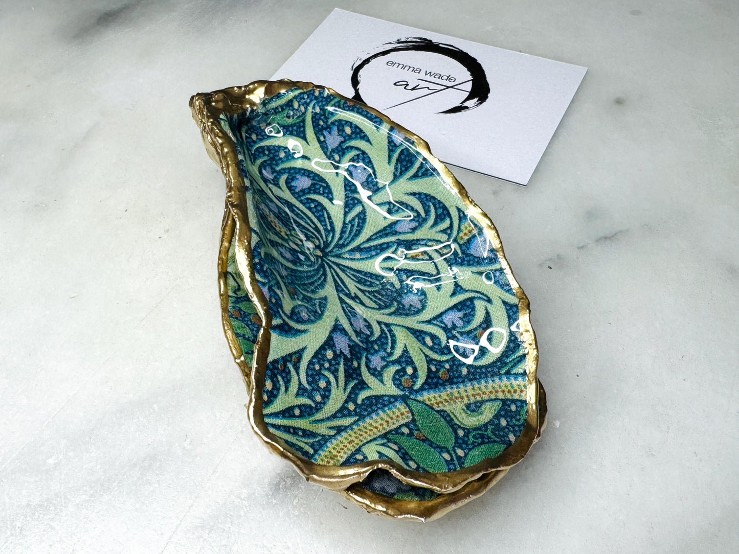 Handmade William Morris inspired oyster shell trinket dish in green and blue tones, finished with gold leaf and sealed with glossy resin. Coastal jewellery dish made from a Dorset oyster shell, featuring a seaweed decoupage design. Perfect for holding rings, earrings and small treasures. Decorative shell bowl photographed on a light background, showing the natural shell shape, detailed pattern and gilded edges.