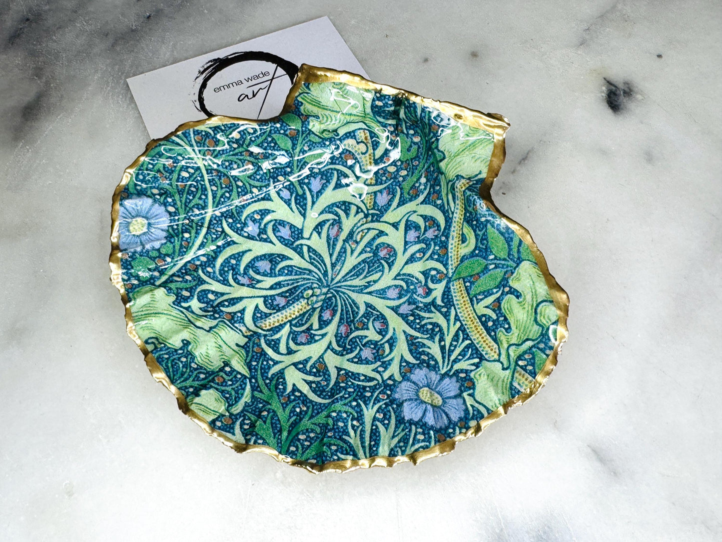 Handmade William Morris inspired scallop shell trinket dish in green and blue tones, featuring a seaweed decoupage design. Finished with gold leaf and sealed with glossy resin. Coastal jewellery dish made from a UK scallop shell, perfect for holding rings, earrings and small treasures. Decorative shell bowl photographed on a light background, showing the natural shell shape and gilded edges.