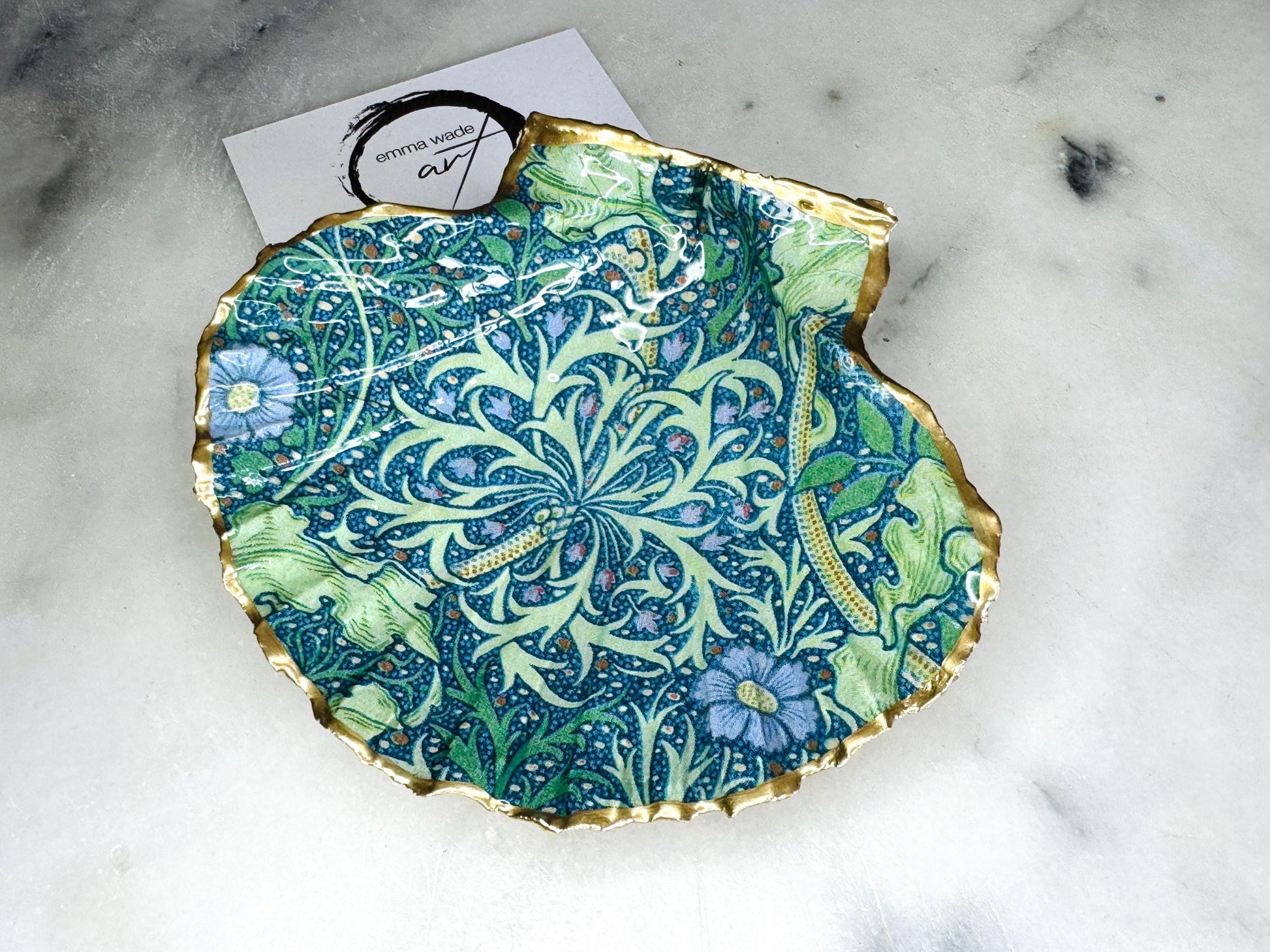 Handmade William Morris inspired scallop shell trinket dish in green and blue tones, featuring a seaweed decoupage design. Finished with gold leaf and sealed with glossy resin. Coastal jewellery dish made from a UK scallop shell, perfect for holding rings, earrings and small treasures. Decorative shell bowl photographed on a light background, showing the natural shell shape and gilded edges.