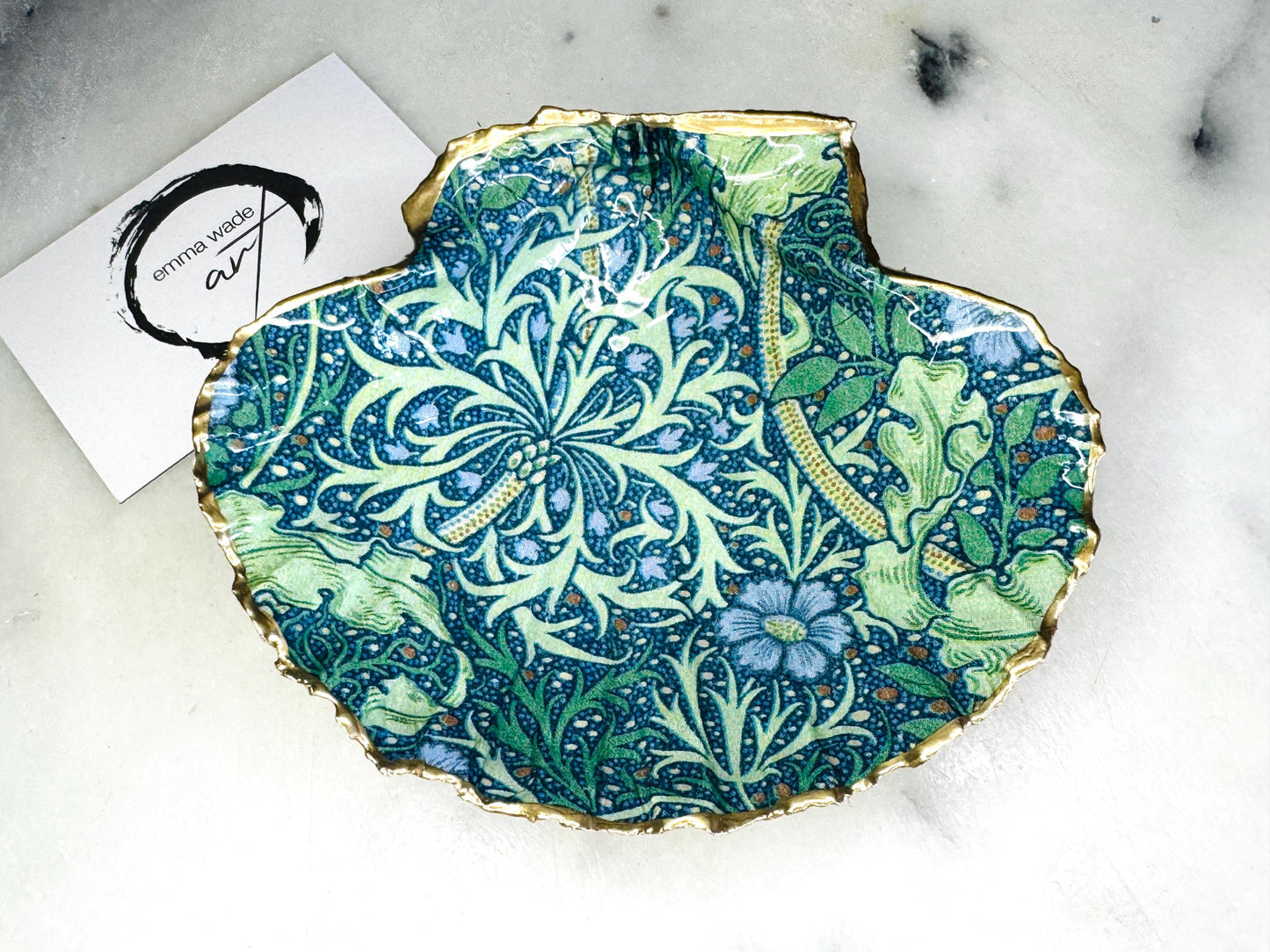 Handmade William Morris inspired scallop shell trinket dish in green and blue tones, featuring a seaweed decoupage design. Finished with gold leaf and sealed with glossy resin. Coastal jewellery dish made from a UK scallop shell, perfect for holding rings, earrings and small treasures. Decorative shell bowl photographed on a light background, showing the natural shell shape and gilded edges.