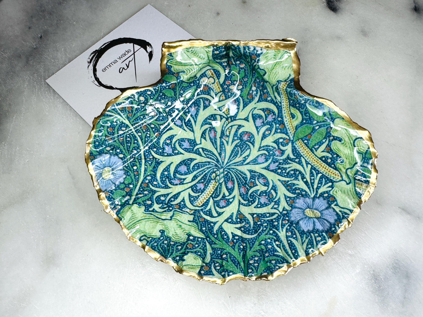 Handmade William Morris inspired scallop shell trinket dish in green and blue tones, featuring a seaweed decoupage design. Finished with gold leaf and sealed with glossy resin. Coastal jewellery dish made from a UK scallop shell, perfect for holding rings, earrings and small treasures. Decorative shell bowl photographed on a light background, showing the natural shell shape and gilded edges.