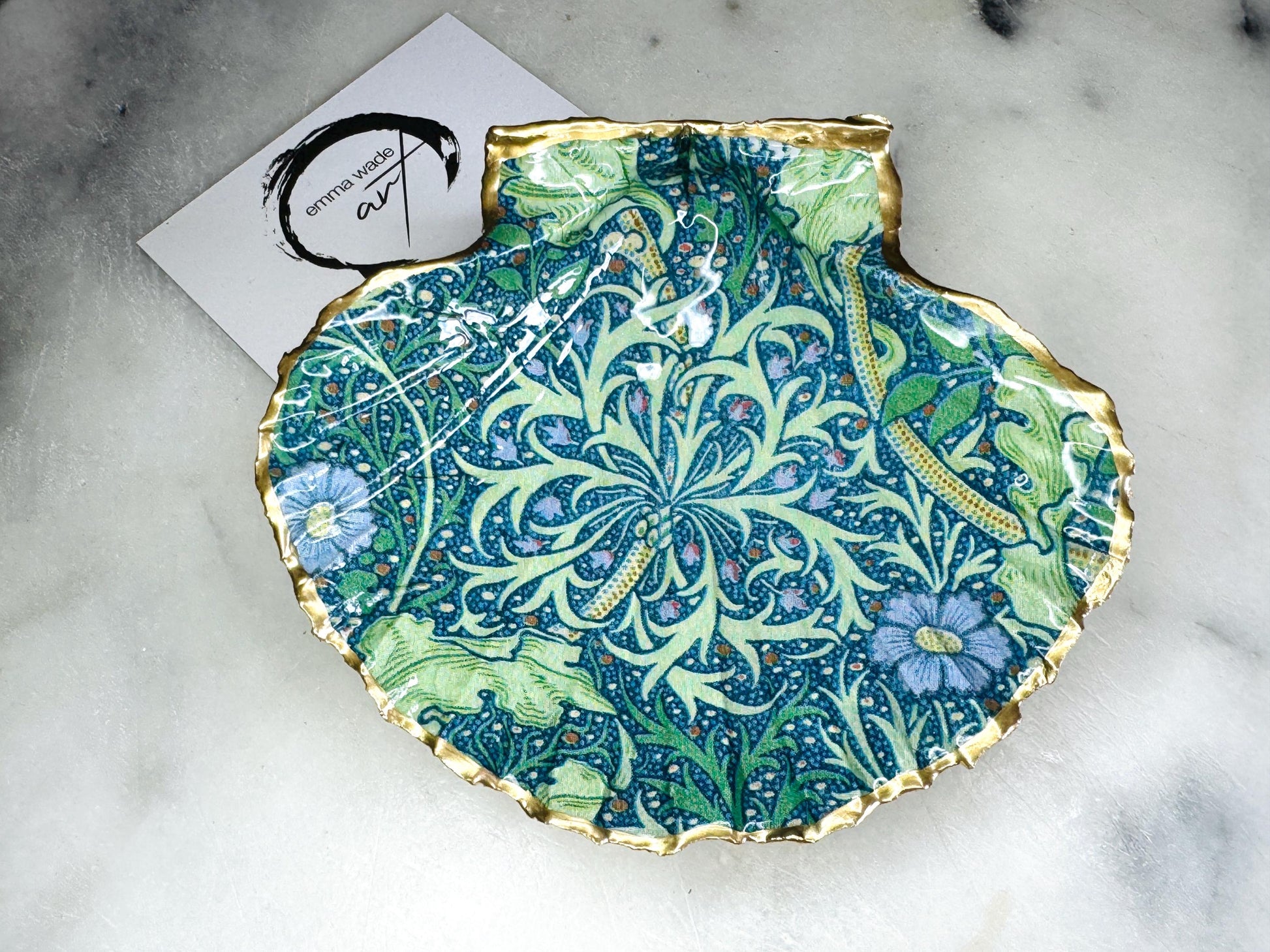 Handmade William Morris inspired scallop shell trinket dish in green and blue tones, featuring a seaweed decoupage design. Finished with gold leaf and sealed with glossy resin. Coastal jewellery dish made from a UK scallop shell, perfect for holding rings, earrings and small treasures. Decorative shell bowl photographed on a light background, showing the natural shell shape and gilded edges.