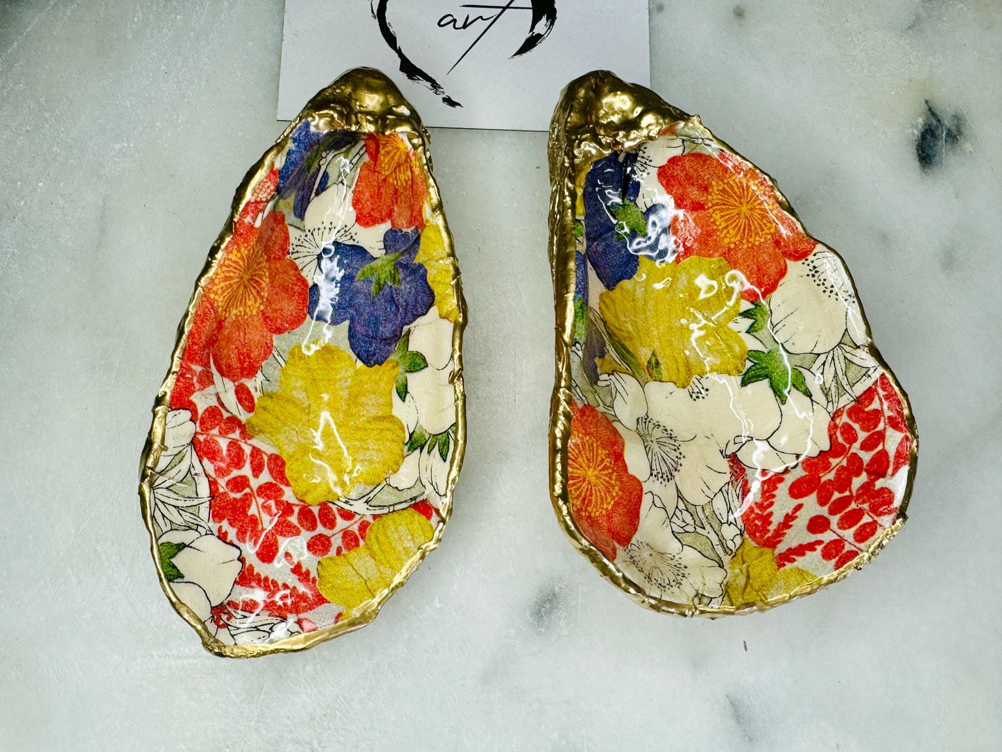 Handmade oyster shell trinket dish featuring a colourful Japanese kimono-style floral design applied with decoupage, finished with hand-gilded gold leaf edges and sealed in glossy clear resin. Natural Dorset oyster shell used as a jewellery dish or ring holder, photographed on a light surface, showcasing the vibrant flowers and luxury gold detailing. Ideal as coastal home decor or a unique handmade gift.