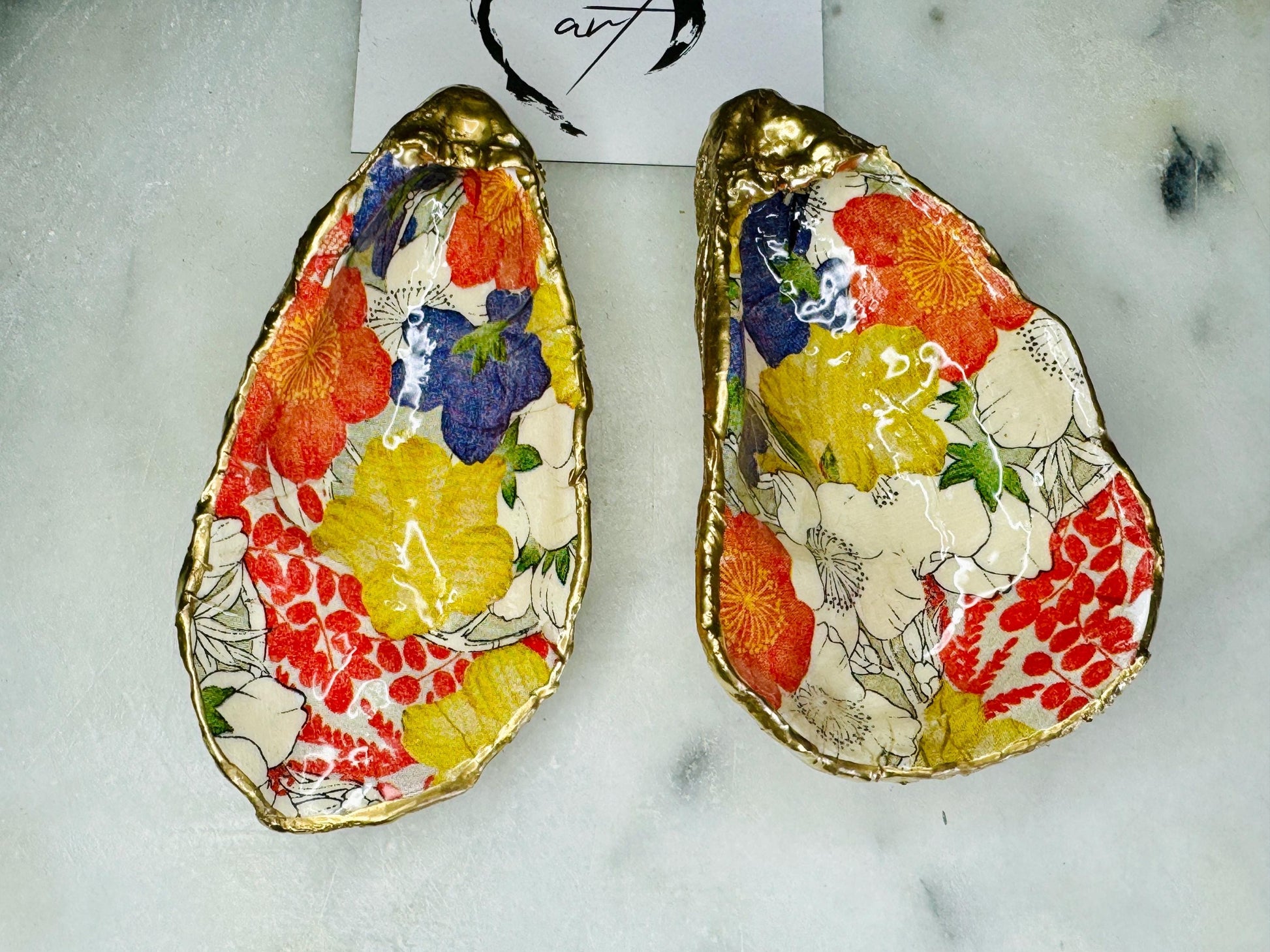 Handmade oyster shell trinket dish featuring a colourful Japanese kimono-style floral design applied with decoupage, finished with hand-gilded gold leaf edges and sealed in glossy clear resin. Natural Dorset oyster shell used as a jewellery dish or ring holder, photographed on a light surface, showcasing the vibrant flowers and luxury gold detailing. Ideal as coastal home decor or a unique handmade gift.