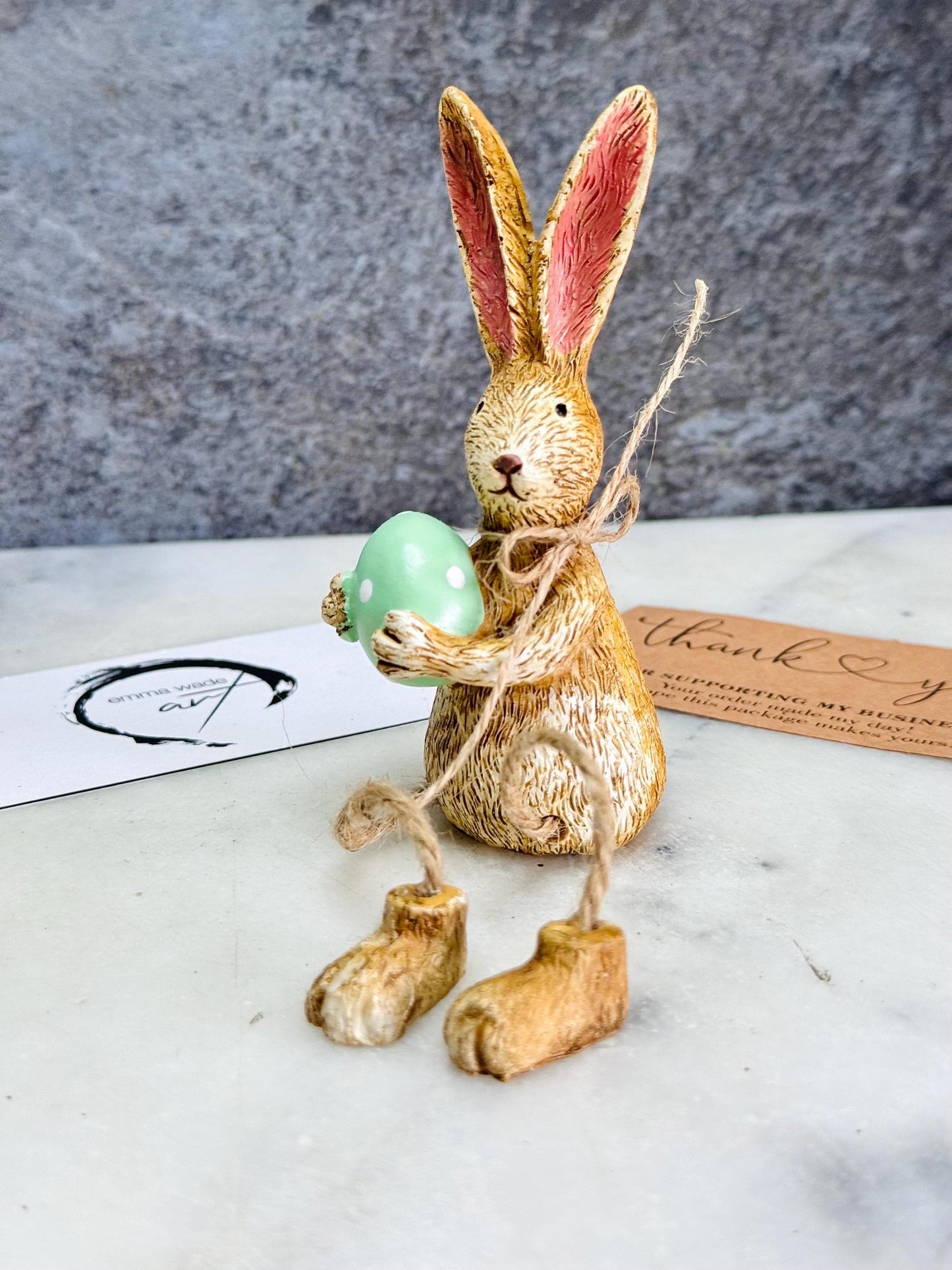 Hand-finished shelf sitting bunny ornament with dangling jute legs and polka dot egg. Available in pink or green. Perfect Easter rabbit shelf decoration, birthday gift, or home décor accessory. Cute animal figurine for rabbit lovers, Easter decorations, new home presents, or seasonal mantel styling.